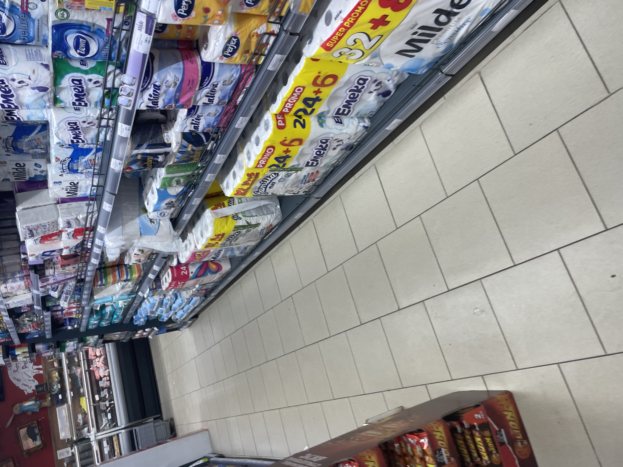 This image captures a wide-angle, slightly tilted view of a well-stocked supermarket aisle in Ihtiman, Bulgaria. The main subjects are towering shelves filled with various brands of toilet paper and paper towels, stretching from the foreground into the background on both sides of the aisle.
The shelves display numerous product packages, predominantly in shades of white, blue, and green, with some yellow and pink accents. Brands visible include "Milde," "Emeka," "Familia," "Deluxe," "EZewa," "Perfex," "Maliva," and "Zebra." Many packages feature promotional banners and text. On the right side, prominent yellow signs declare "PROMO" and "SUPER PROMO," highlighting deals such as "24+6" (likely indicating 24 rolls plus 6 free) and "32+" for larger packs. One package of "Milde" is marked "Sensitive" and "Premium." Text like "Delicate Care" and "PLUS" is also visible on other products.
The floor of the aisle is covered in light-colored, square tiles with dark grout lines, leading the eye towards the back of the store. In the bottom right corner of the foreground, a cardboard display stand with "LION" branding, presumably for chocolate bars, is partially visible. Further down the aisle on the left, other retail displays and what appears to be a refrigerated unit can be seen. The scene is brightly lit, typical of an indoor retail setting. No individuals are present in the image.