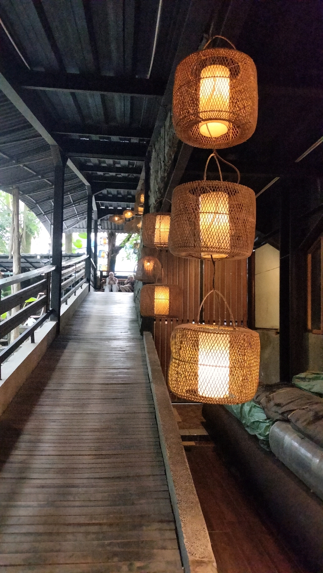 This is a dimly lit outdoor walkway with a rustic aesthetic, likely located in the city of Berastagi, Indonesia. The walkway is made of weathered wooden planks, extending from the foreground into the distance where several people are seated. On the right side of the walkway, a series of woven rattan pendant lamps hang from the ceiling, casting a warm, golden glow. These lamps are cylindrical in shape with an open, criss-cross weave, revealing the light bulbs within. The ceiling above the walkway is constructed of dark wooden beams and corrugated metal roofing. The left side of the walkway features a wooden railing. In the background, trees and foliage are visible, suggesting a natural or semi-natural setting. The overall impression is one of a serene and inviting pathway.