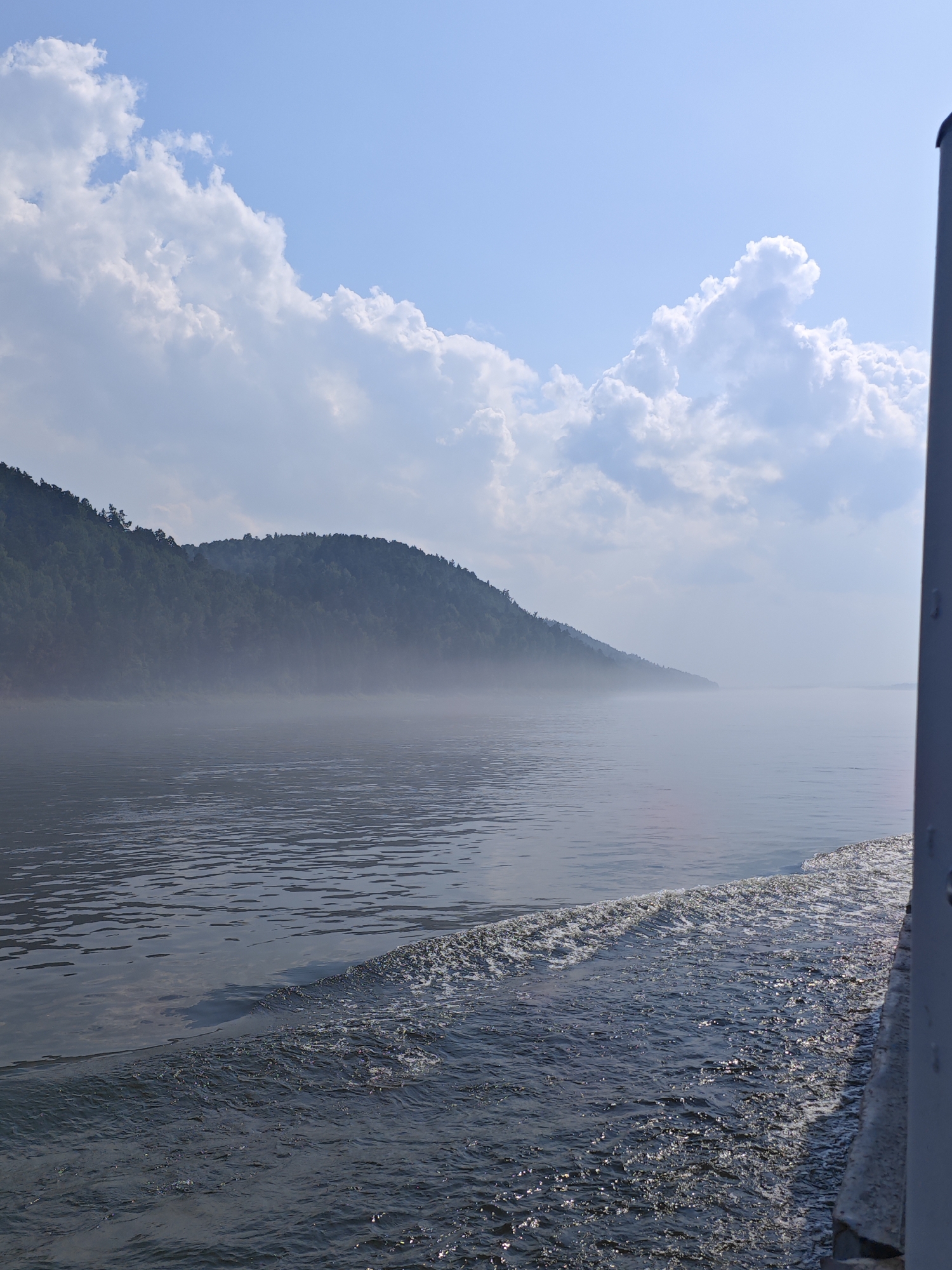 A view from a moving boat on a wide river. The river surface is rippled, with a visible wake trailing from the boat on the right side of the frame. On the left, a dense, tree-covered hillside rises steeply from the water's edge. A light mist hangs low over the water and at the base of the hill, obscuring the distant shoreline. The sky above is a clear blue with scattered white, puffy cumulus clouds. The image captures a serene, natural landscape. The location is the Yenisei River near the city of Predivinsk, Russia.