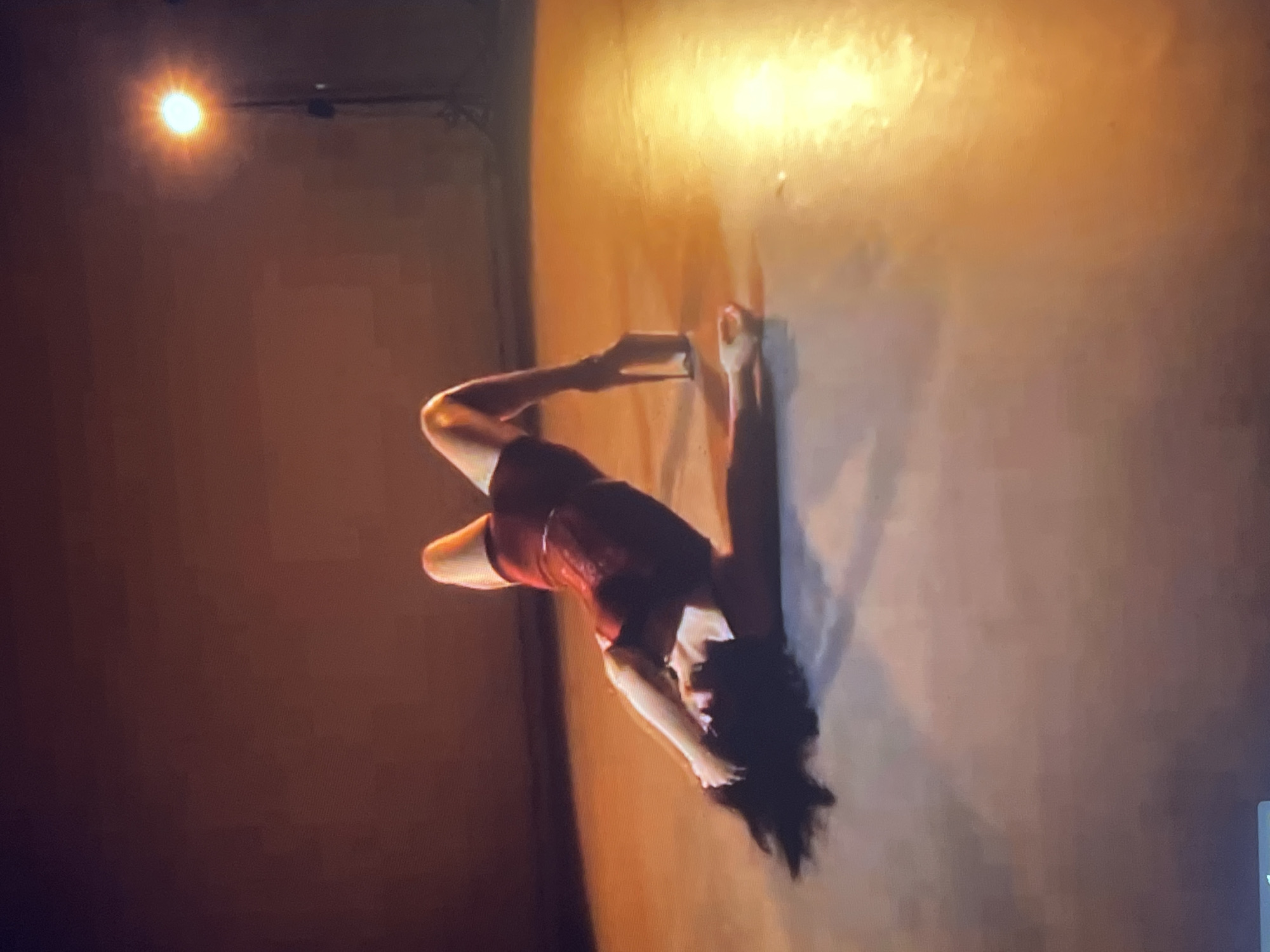 A person with dark hair, wearing a red outfit and extremely high-heeled shoes, is lying on their back on a light-colored floor with their hips arched upwards. Their arms are behind their head, and their legs are bent. The background is a dark, reddish-brown color, with a bright light source on a stand in the upper right corner, casting a warm glow on the scene. The overall impression is that of a performance or artistic pose. The scene does not provide any specific information about the city of Zoriane, Ukraine.