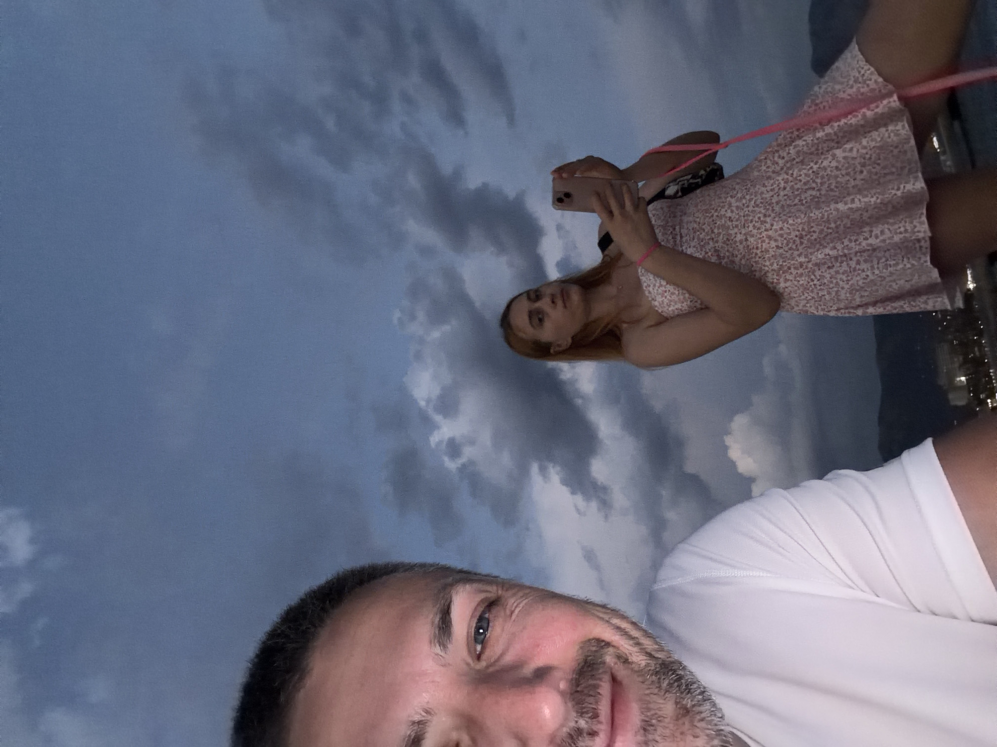 In the foreground, a man with short graying hair and a stubbled beard smiles at the camera, his left eye visible. He wears a white t-shirt. Behind him, a young woman with long blonde hair, wearing a strapless pink floral print dress, holds up a pink smartphone, taking a picture or video. She has a black bag slung over her shoulder and wears pink bracelets on both wrists. The background is dominated by a dramatic, cloudy sky, with a hint of a city skyline and mountains visible in the distance, suggesting a coastal urban setting like Budva, Montenegro, possibly at dusk or during inclement weather.