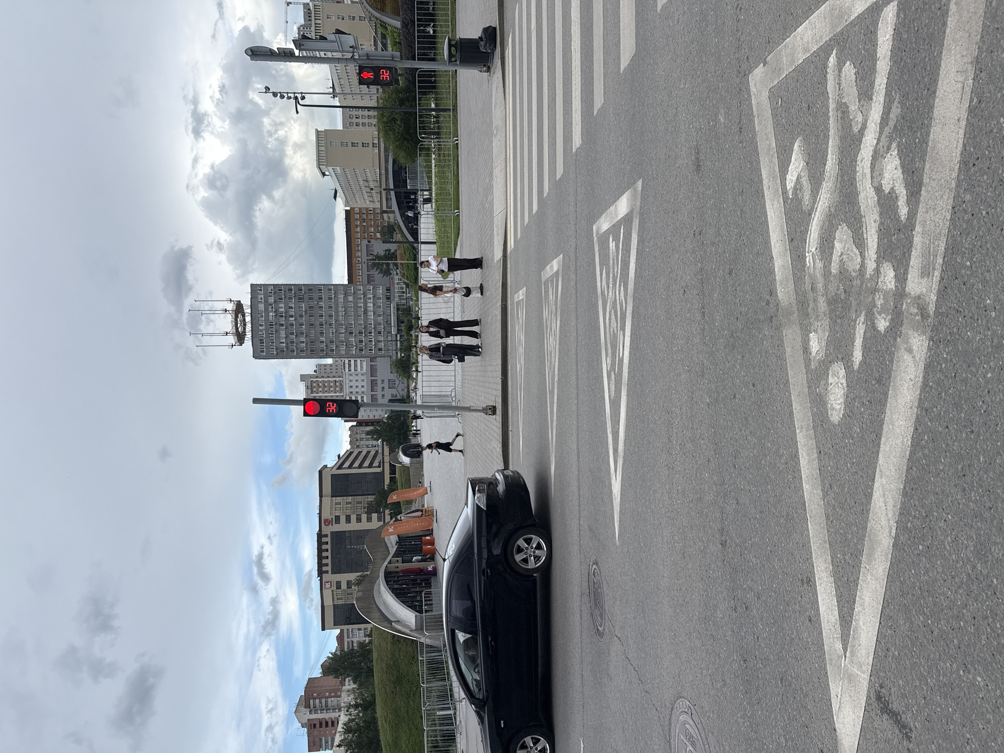 This is a street-level, full-shot, portrait-orientation photograph taken in Perm, Russia. The scene depicts a crosswalk with white markings, including a pedestrian signal showing a red "walk" symbol and a countdown timer displaying "32". Several people are visible on the sidewalk near the crosswalk, some appearing to wait for the signal to change. A black car is parked or stopped on the right side of the road, aligned with the crosswalk. In the background, modern buildings and a cloudy sky suggest an urban environment. The overall lighting indicates it is daytime, possibly late morning or afternoon, with overcast conditions contributing to the diffused light. The image captures a typical urban moment at a street intersection.