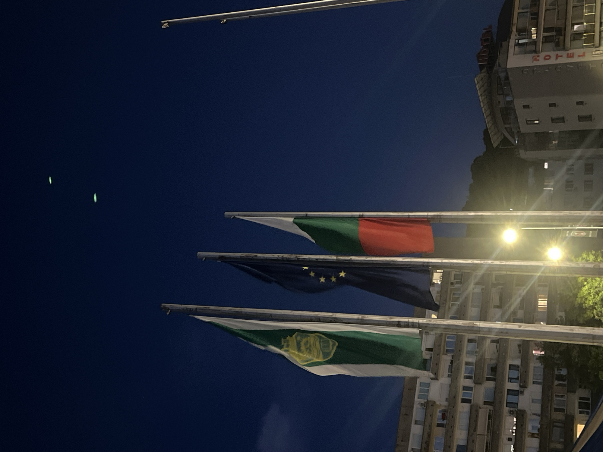 In the city of Unknown, Unknown, at night, three flags are displayed on flagpoles in front of a building complex. The flag on the left is green and white with a crest in the center. The middle flag is dark blue with yellow stars, resembling the European Union flag. The flag on the right is a tricolor of red, white, and green. Behind the flags, several multi-story buildings are visible, with some windows illuminated. Streetlights are also present, casting a warm glow. In the dark night sky, three faint, elongated green lights are visible. The overall scene depicts an urban setting at night with national and international symbols prominently displayed.
