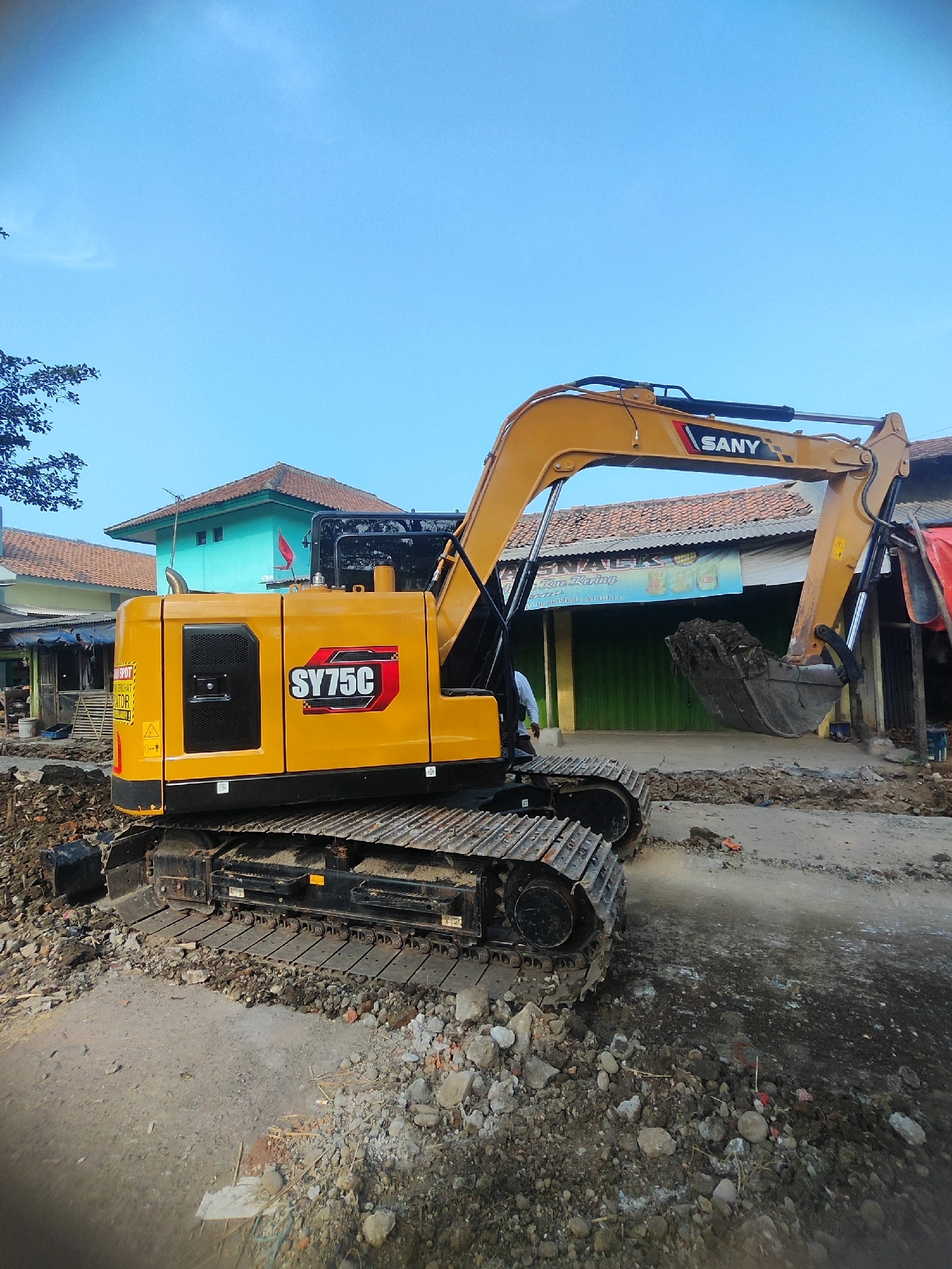 In Karawang, Indonesia, a yellow SANY SY75C excavator is positioned on a dirt and debris-strewn ground in what appears to be a construction or demolition site. The excavator's tracks are resting on uneven terrain mixed with rocks and soil. The excavator's boom is raised, and its bucket is filled with dirt, suggesting it has been actively digging or moving material.
In the background, there are buildings with terracotta-tiled roofs and light blue walls. One building features a green corrugated metal facade. A sign with the word "SNACKS" is visible on the building with the green facade, along with text in Indonesian that appears to be a business name. To the left of the excavator, some trees with dark green foliage are present. The sky above is a clear, light blue. A single individual, wearing a light-colored shirt and dark pants, is standing to the left of the excavator, seemingly observing the work. The overall environment suggests an outdoor setting in a populated area, likely a roadside or commercial strip undergoing some form of excavation or reconstruction.