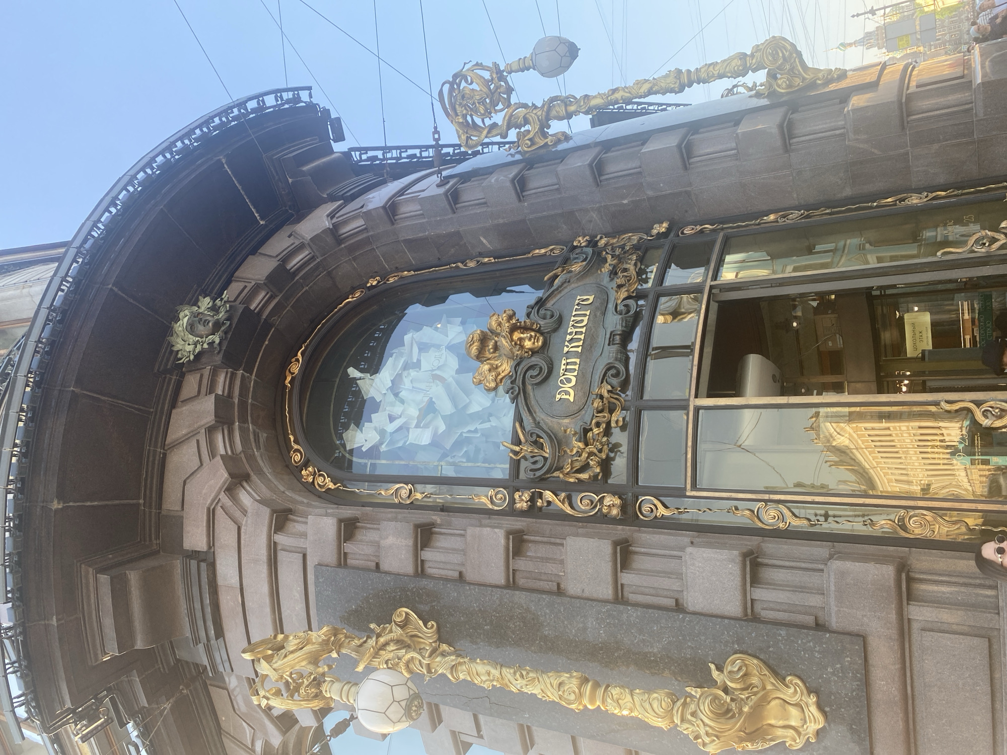 The image shows the exterior of the "House of Books" (Dom Knigi) building in Saint Petersburg, Russia. The architectural details include ornate gold embellishments around the windows, a dark stone facade, and decorative sculptures. The signage, "Дом Книги," is visible on the window. A large window is filled with white objects. The sky is visible in the background.