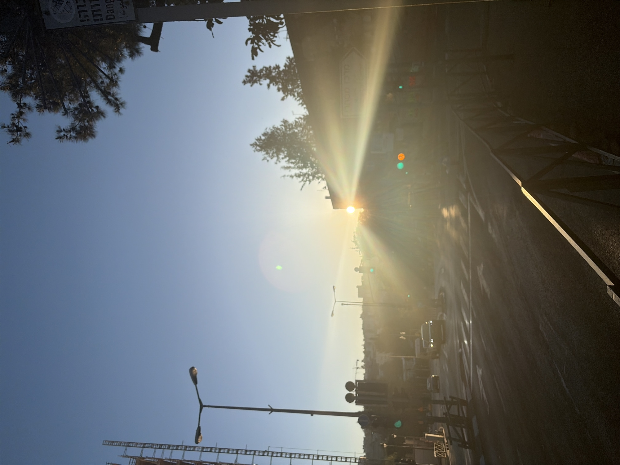 The image depicts a street in Jerusalem, Israel, captured during sunrise or sunset. The sun is positioned at the end of the street, casting a strong glare and lens flare that obscure some details. Streetlights and traffic signals are visible, indicating a city environment. Construction scaffolding can be seen in the distance, along with trees lining the street. The road itself is black with white lines, suggesting it is paved. A partial view of a guardrail or barrier is visible on one side. The overall impression is of a city street scene during a time of bright sunlight.