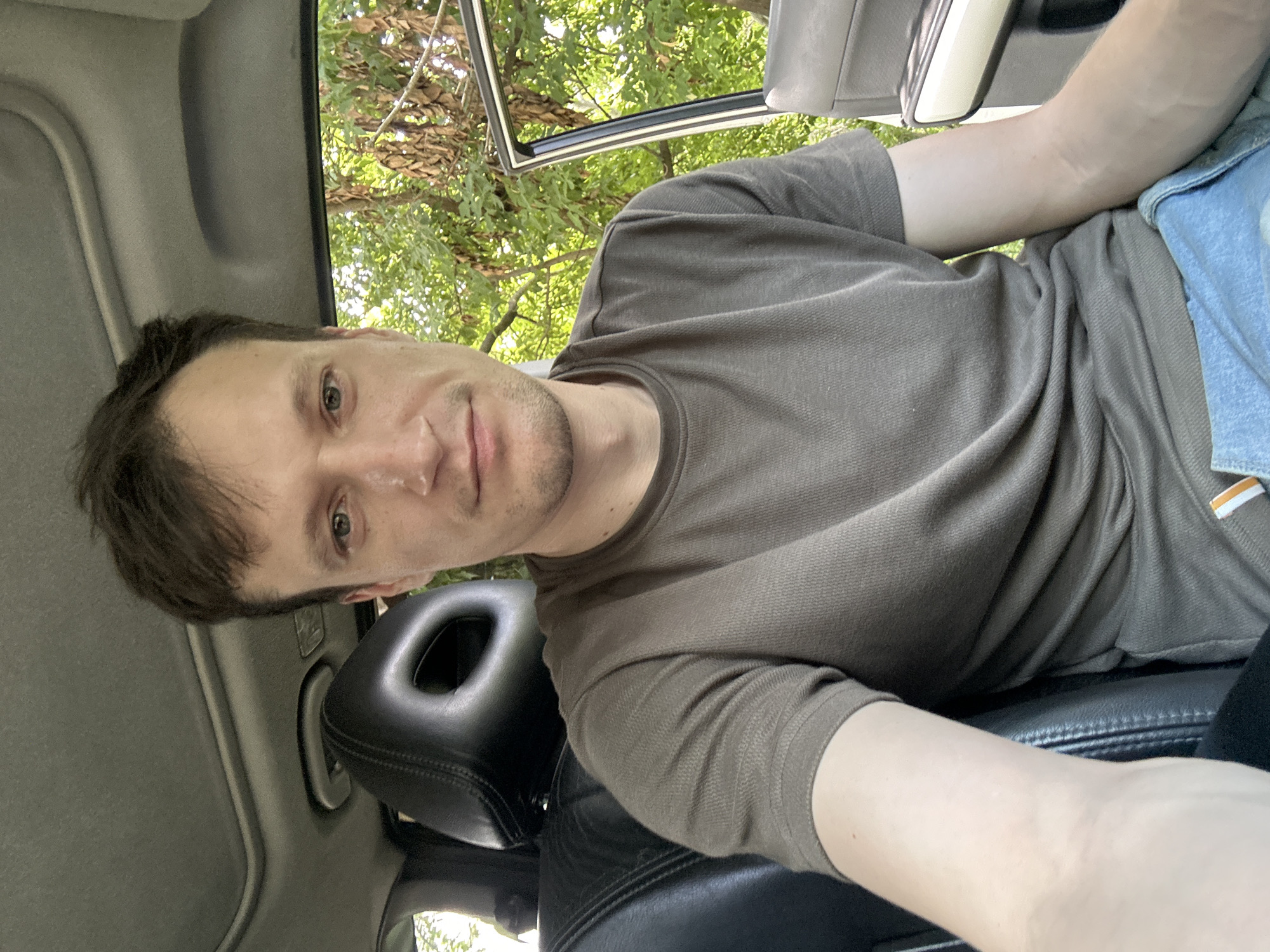 A man is sitting in the driver's seat of a car. He is wearing a green t-shirt and blue jeans. The car appears to be parked outdoors, with trees and foliage visible through the open passenger door and window. The interior of the car features black leather seats and a sunroof. The scene is consistent with a location in or around Makiv, Ukraine, given the foliage and general appearance.