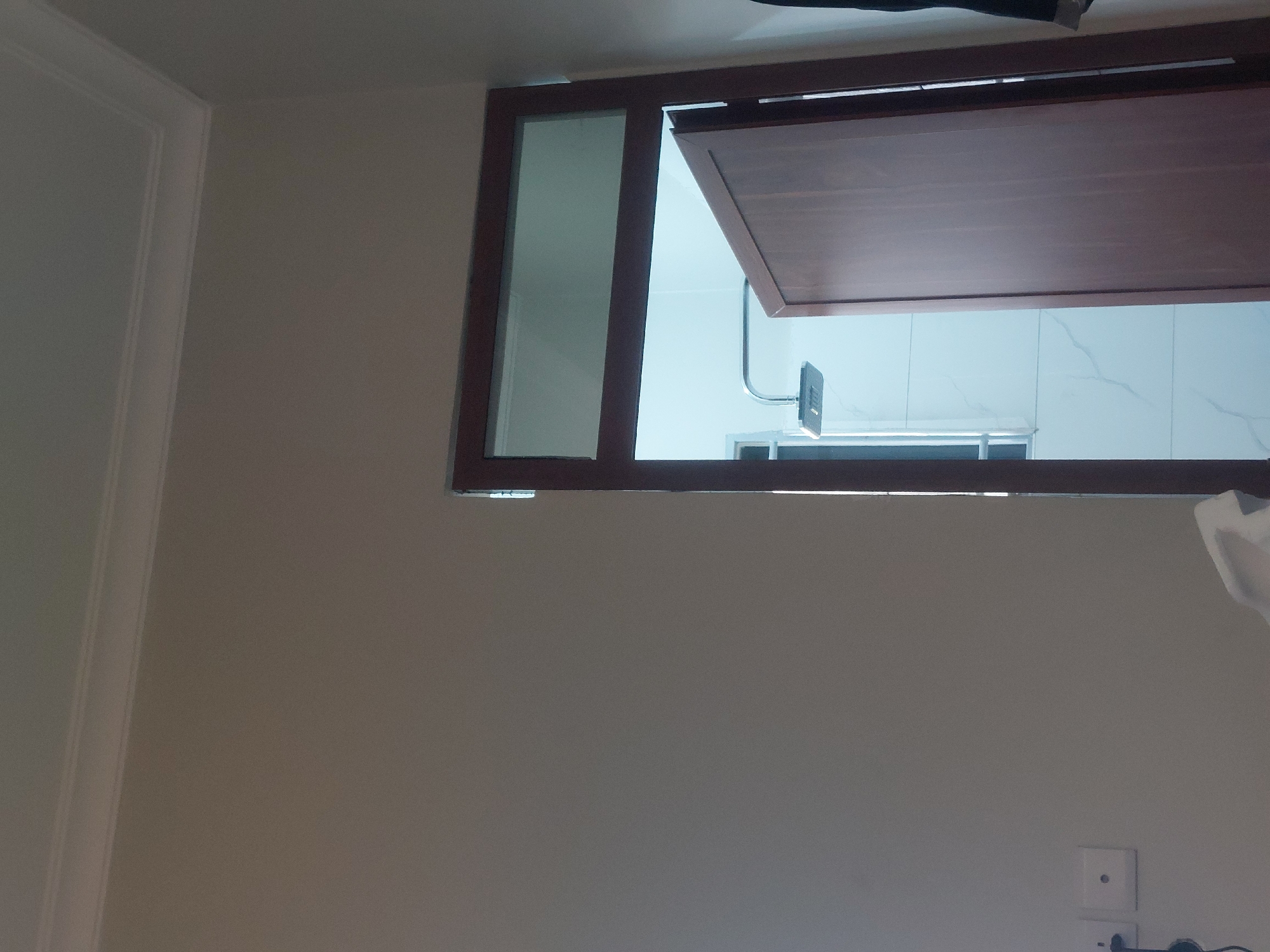 The image shows a partially opened bathroom window. The window frame is dark brown, with a clear glass panel on the left and a hinged wooden panel on the right. Through the window, a section of the bathroom is visible, including a shower head, a partial view of what appears to be a shower bench, and white marble-patterned wall tiles. The wall surrounding the window is a light, warm gray. In the lower right corner, an electrical outlet is visible on the wall. The location appears to be indoors. It is impossible to confirm the city without further information.