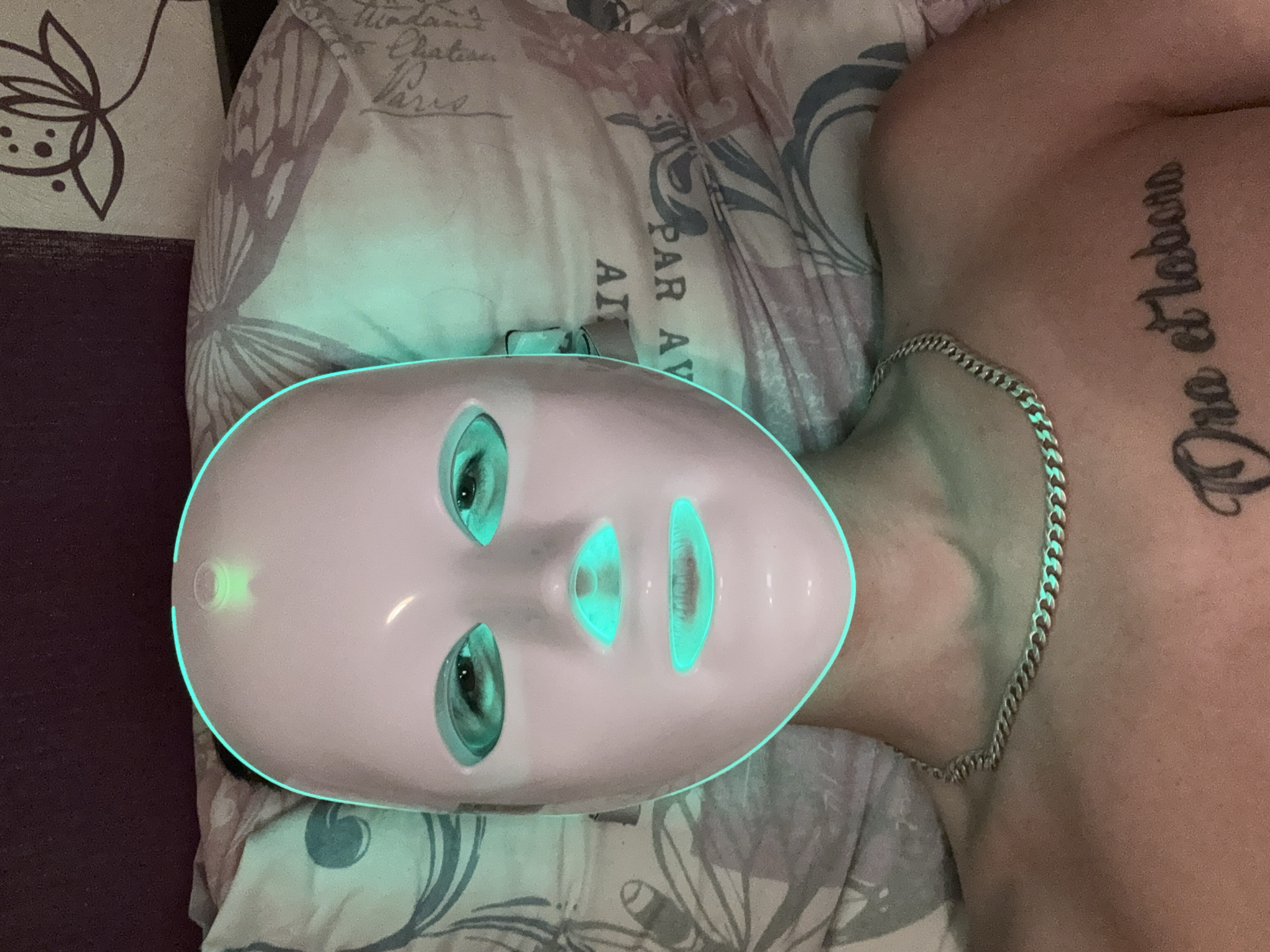 A person is lying down, wearing a pink LED facial mask with green lights emanating from the eye, nose, and mouth areas. The mask covers their face from the forehead to below the chin. They have a silver chain necklace on their chest, and a tattoo with the Latin phrase "Ora et labora" is visible on their upper chest. The person is on a bed with a patterned pillowcase, which has floral and butterfly motifs. A dark purple headboard is visible in the background. The overall setting suggests an indoor domestic environment. There is no discernible information in the image to suggest a location in Stara Zagora, Bulgaria.