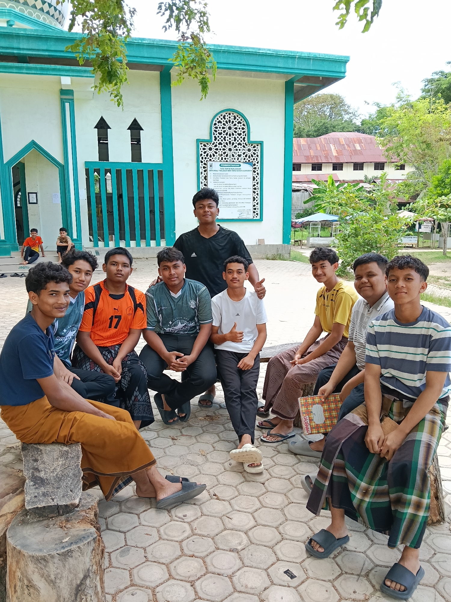 A group of nine young men are gathered outdoors, likely near a mosque in Lhokseumawe, Indonesia. The men are dressed in a mix of casual attire: t-shirts, jeans, and sarongs. They are sitting and squatting on a paved surface. A mosque with a turquoise and white color scheme is visible in the background. Several individuals are wearing sandals. A building with a red roof can be seen in the distance. The scene appears to be a casual gathering of young men.