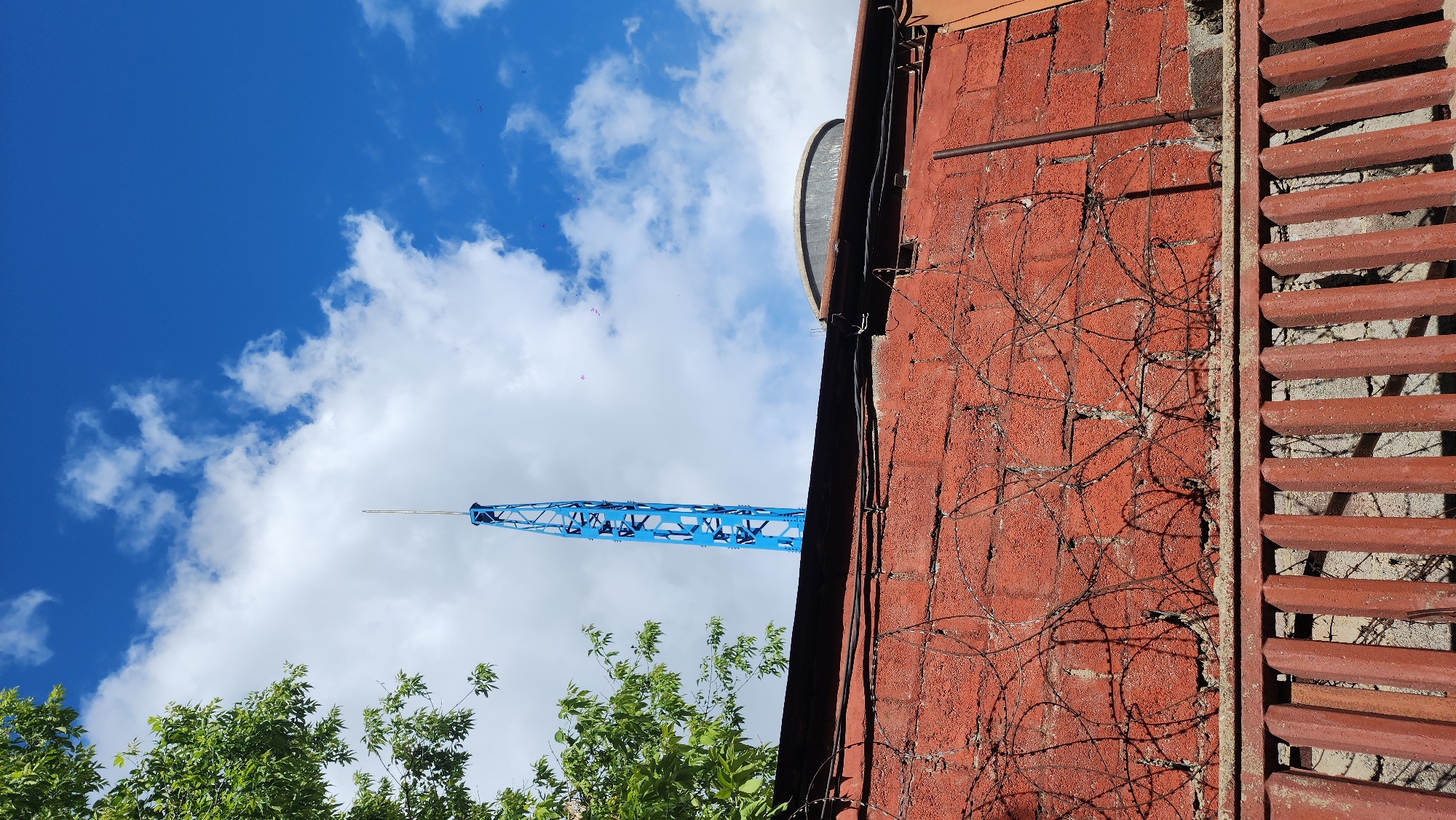 A low-angle shot shows a red brick wall with a ventilation opening and barbed wire, framing a view of a blue tower crane extending into a blue sky with cumulus clouds. Green foliage from trees is visible at the bottom left. The crane and foliage suggest a construction site or urban development zone. This is the Ostankino Tower in Moscow, Russia. The tower is visible in the image extending to the sky, near the clouds.