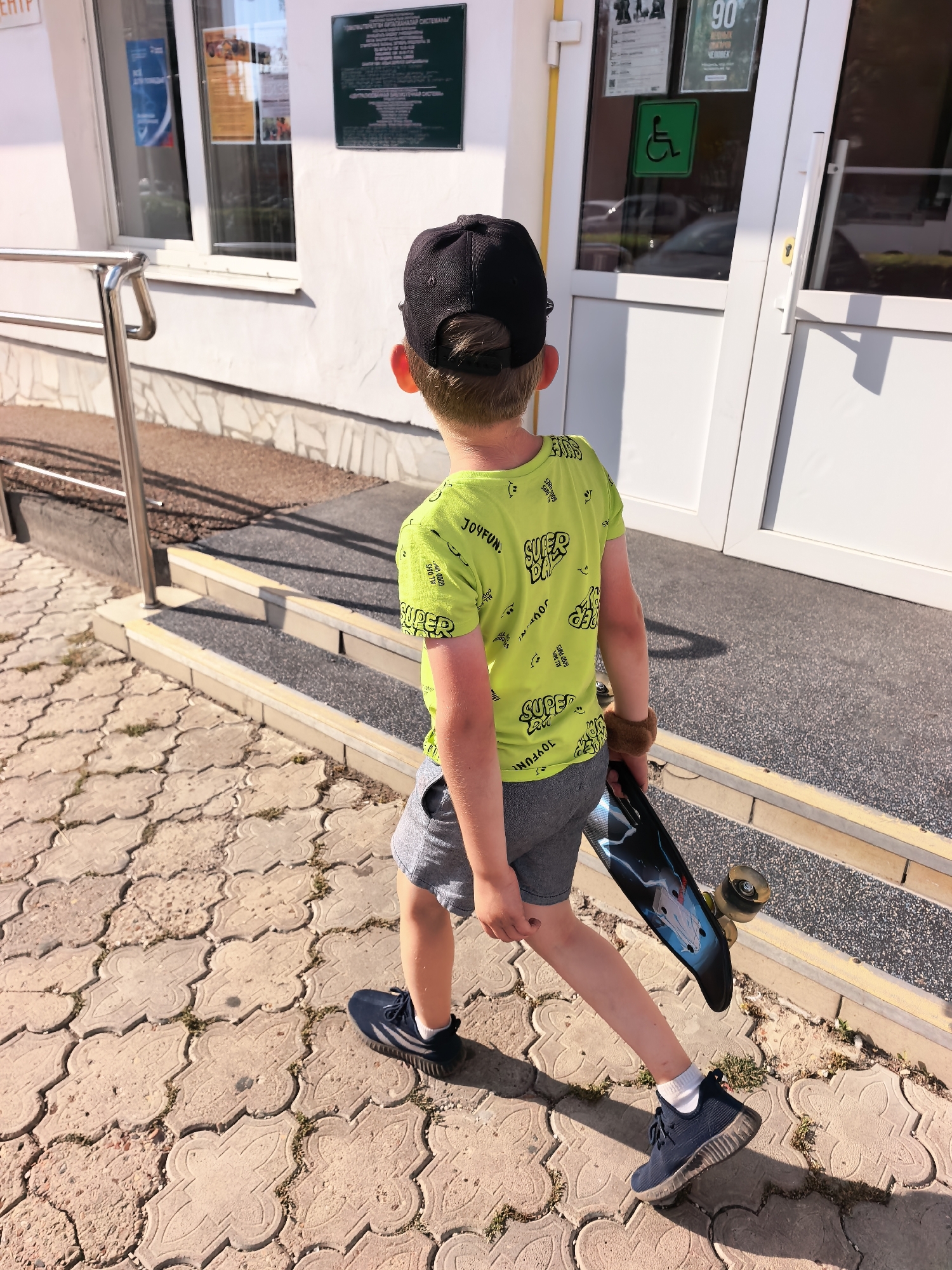A young boy wearing a black baseball cap, a neon green t-shirt with a "Super Dude" graphic print, and grey shorts walks away from the camera carrying a skateboard. He is on a patterned concrete sidewalk in front of a white building with large glass doors and windows. To the left of the building is a metal handrail leading to a ramp with steps. The building has a green sign with text in Russian and a smaller green sign with a wheelchair accessibility symbol on the door. The scene appears to be outdoors on a sunny day. The location is identified as Sterlitamak, Russia.