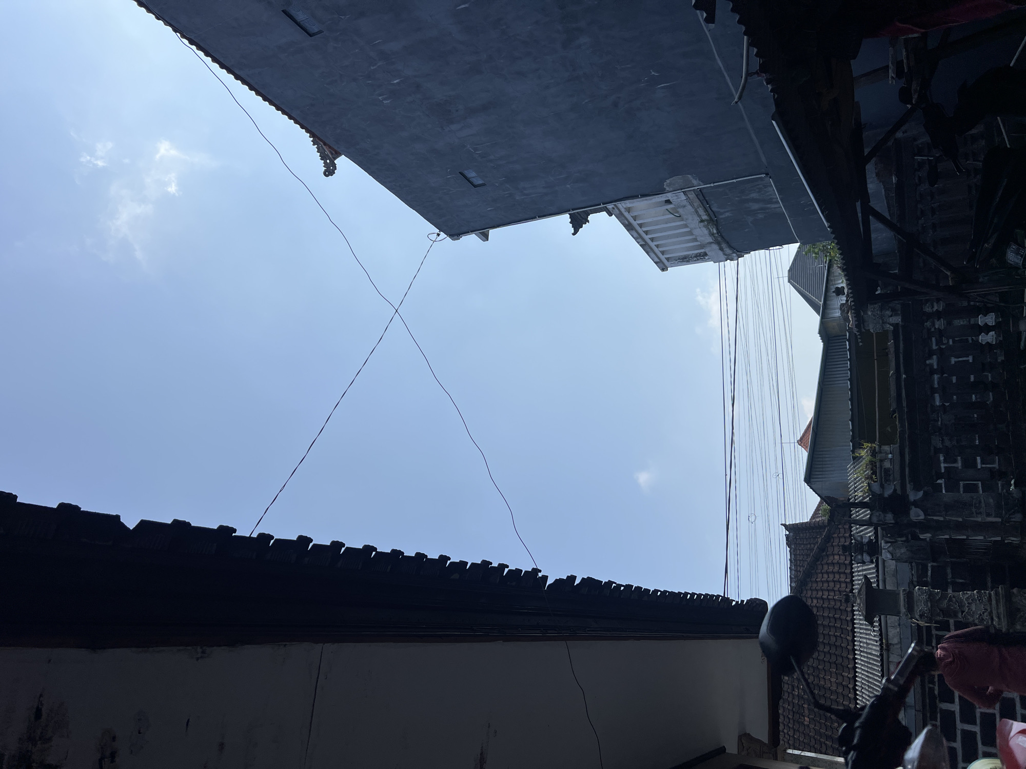 The image captures a view of the sky and rooftops in Denpasar, Indonesia. Two buildings are prominent in the foreground, one on the left with a dark, overhanging roof, and another on the right with a dark grey facade. Overhead, a bright blue sky is visible with a few wispy clouds. Electrical wires crisscross the sky between the buildings. In the background, multiple rooftops of residential buildings, some with tiled roofs and others with corrugated metal, are discernible. A stone wall or fence, likely part of a property boundary, runs horizontally across the mid-ground. Partially visible at the bottom left is a portion of a motorcycle, including its handlebar and mirror. To the left of the stone wall, a person wearing a reddish-brown hooded jacket is standing and facing away from the camera. The scene suggests an urban or semi-urban environment in Denpasar, likely a residential area, under a clear daytime sky.