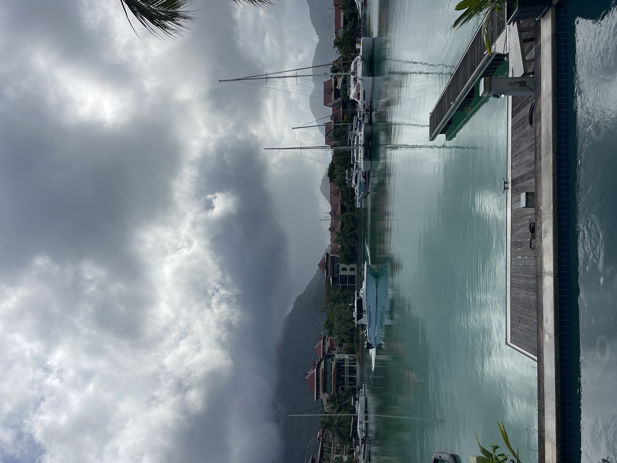 The image shows a marina on a cloudy day. In the foreground, there is a swimming pool with a tiled edge, and a wooden deck with a long pier extending into the water. The water is a clear turquoise color and reflects the sky and surrounding buildings. In the middle ground, several boats are docked, including sailboats and a motorboat that is underway, leaving a wake. Behind the boats, there are multi-story buildings with red-tiled roofs, surrounded by palm trees. In the background, lush green mountains rise up, partially obscured by heavy, dark clouds. The scene depicts a harbor in Providence, Seychelles, under overcast and potentially stormy weather.
