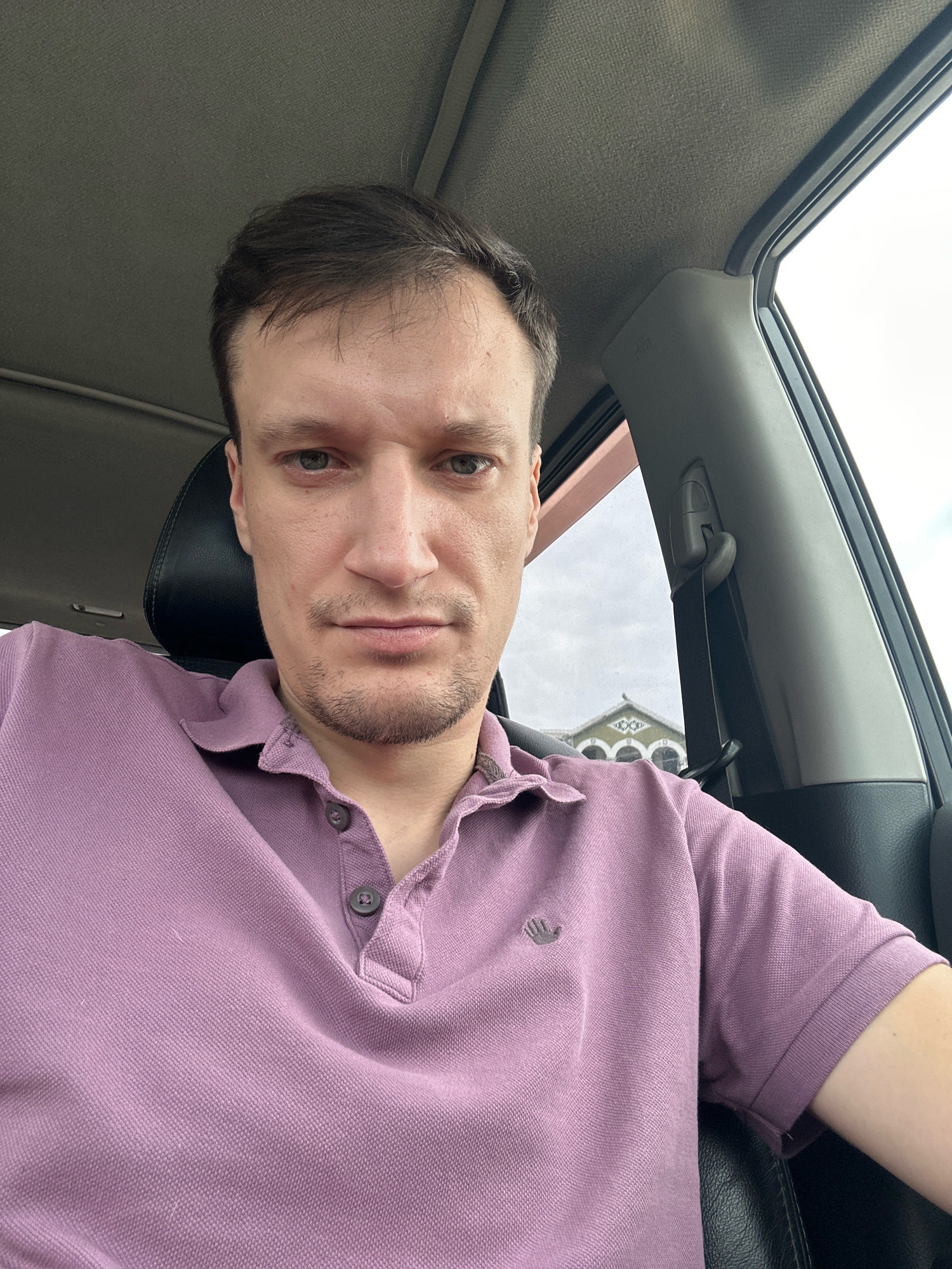 A close-up, over-the-shoulder selfie shows a man with short brown hair, blue eyes, and a light beard sitting in the driver's seat of a car. He is wearing a light purple polo shirt. The car's interior is visible, including a black headrest, part of the dashboard, and a seatbelt. Outside the car window, a building with arched windows and a decorative facade is visible against a cloudy sky. The location is identified as Shatava, Ukraine.