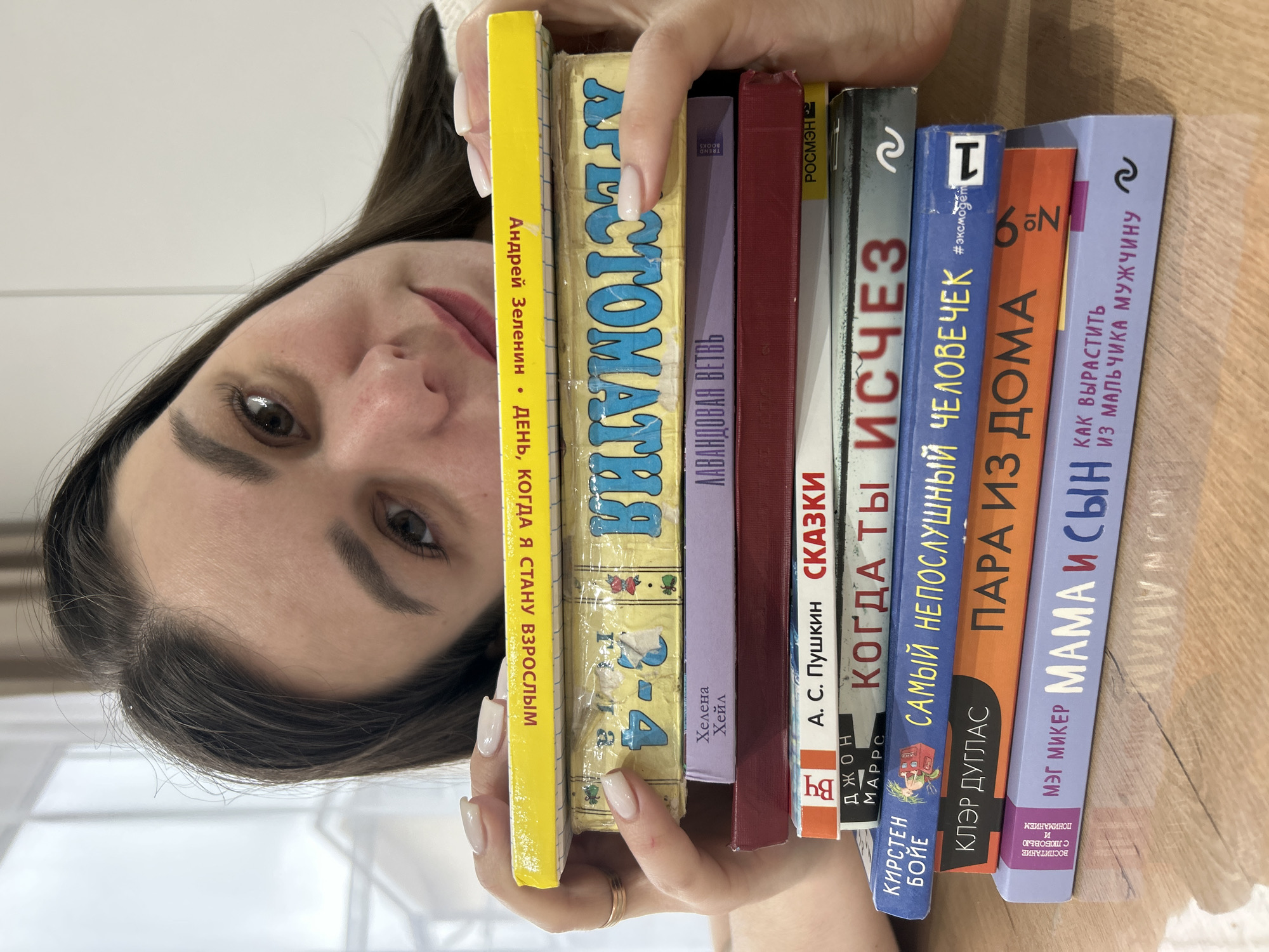 A close-up, eye-level photograph captures a person holding a stack of ten books, with only the upper part of their face and hands visible. The person has dark hair, a neutral expression, and is looking towards the camera. They have manicured nails with light-colored polish and a simple gold-toned ring on one finger. The books are held horizontally, with their spines facing the viewer, suggesting a curated collection.
The books, written in Russian, feature various titles and authors. From left to right, they include: "День, когда я стану взрослым" by Андрей Зеленин, an "Агестоматия" (possibly "Антология") volume 2-4, "Лавандовая ветвь" by Хелена Хейл (Helena Hael), "Сказки" by А. С. Пушкин (A. S. Pushkin), "Когда ты исчез" by Джон Маррс (John Marrs), "Самый непослушный человечек" by Кирстен Бойе (Kirsten Boye), "Пара из дома" by Клэр Дуглас (Claire Douglas), and "Мама и сын" by Мэг Микер (Meg Meeker), subtitled "Как вырастить из мальчика мужчину" (How to raise a boy into a man) and "Воспитание с любовью и пониманием" (Raising with love and understanding). Publishers like "Росмэн" (ROSMEN) and "Trend Books" are also visible.
The scene appears to be indoors, with a light-colored background and surface, likely a table, on which the books rest. The person's face is oriented upside down relative to the books, implying a unique perspective or pose for the photograph. The image portrays a moment of showcasing literature.