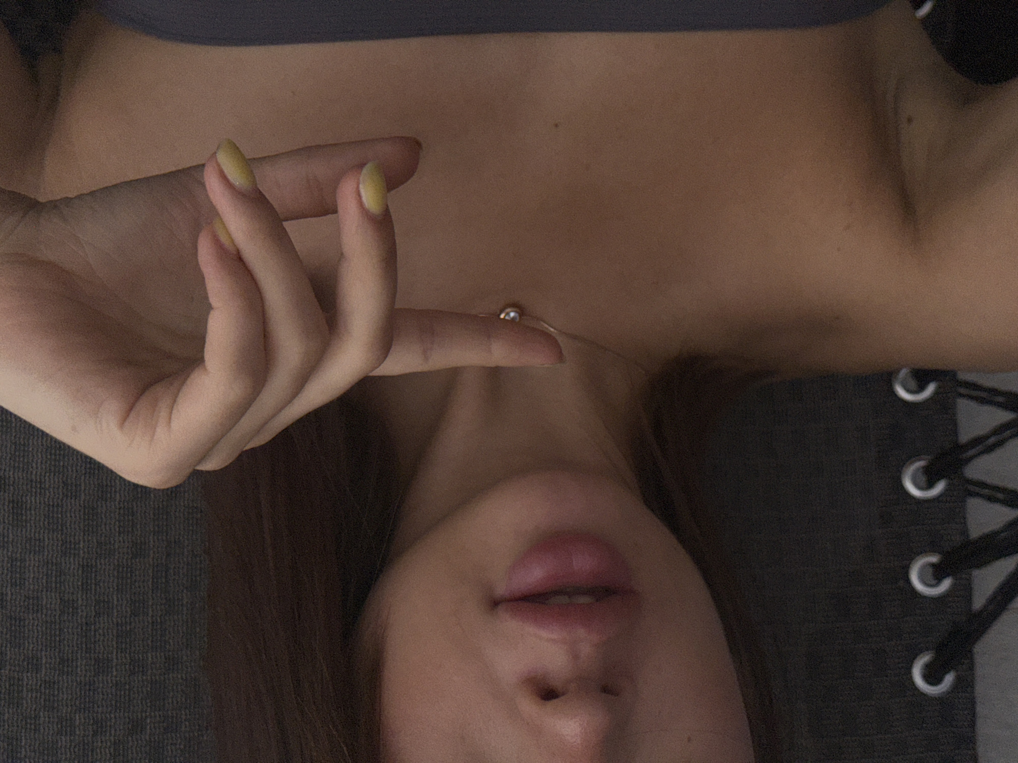 The image shows a close-up of a young woman's face and upper body. She has dark hair, full lips, and is wearing a strapless top. A delicate necklace with a small pendant is visible around her neck. Her hand, with yellow nail polish, is raised and touching her chest near the necklace. The background appears to be a textured, dark fabric, possibly part of a chair or seating. The lighting is soft and diffused, suggesting an indoor setting. There is no information in the image that would indicate a location in Lobanovo, Russia.