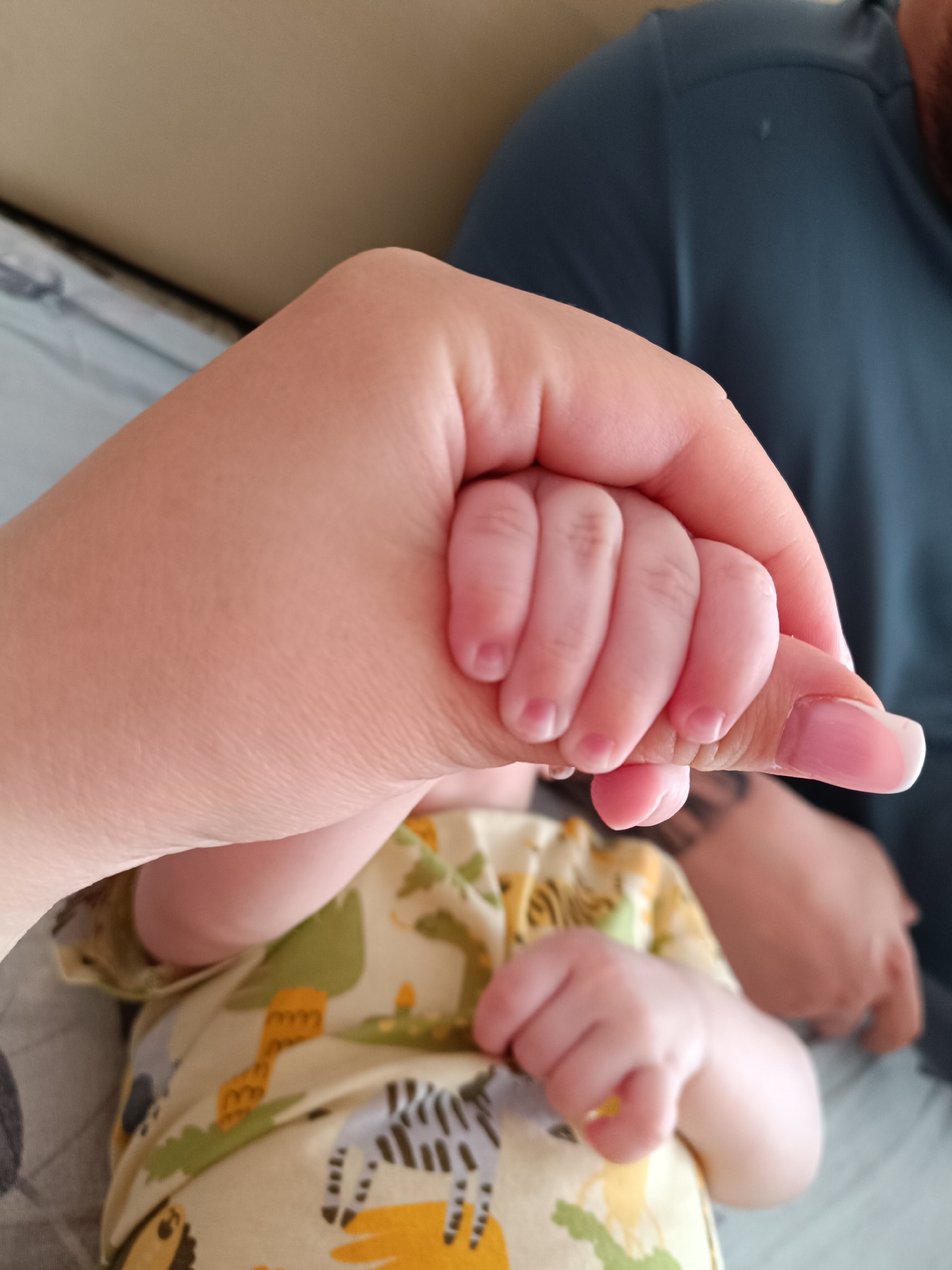 A close-up shot focuses on a baby's hand tightly gripping an adult's index finger. The baby is wearing a light-colored onesie adorned with animal prints. A portion of another adult can be seen blurred in the background, wearing a dark-colored shirt. The environment appears to be indoors, possibly a home setting. The location is not identifiable as Tobolsk, Russia based on the visual information.