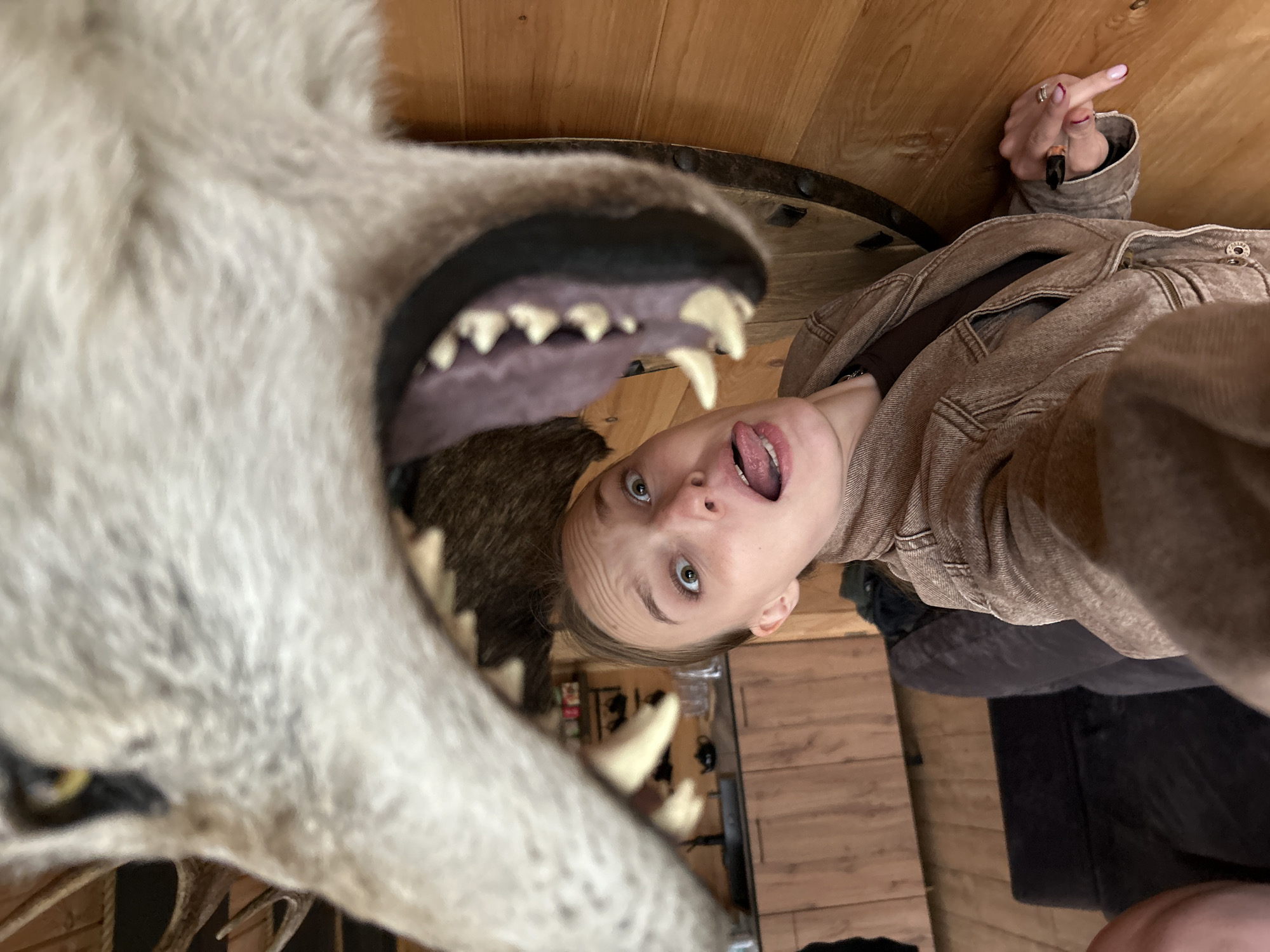 The image is a portrait of a young woman from a low-angle perspective. She is looking directly at the camera with her mouth open and tongue sticking out, her eyes wide open. Her expression appears playful or surprised. She is wearing a light brown denim jacket over a dark brown top. On her left hand, she wears a silver ring. Her right hand is raised, and she is making a gesture with her middle finger extended. The scene is indoors, with wooden paneling on the walls and floor. To the left of the woman is a taxidermied wolf's head with its mouth open, revealing sharp teeth. The wolf's fur is light gray. The overall lighting suggests an indoor setting, possibly with artificial light. The image seems to be a selfie taken in a room that may have rustic decor, perhaps a cabin or a themed restaurant, given the presence of the taxidermied animal. The woman's gesture and expression indicate a lighthearted and possibly cheeky mood. There is no visible text in the image.