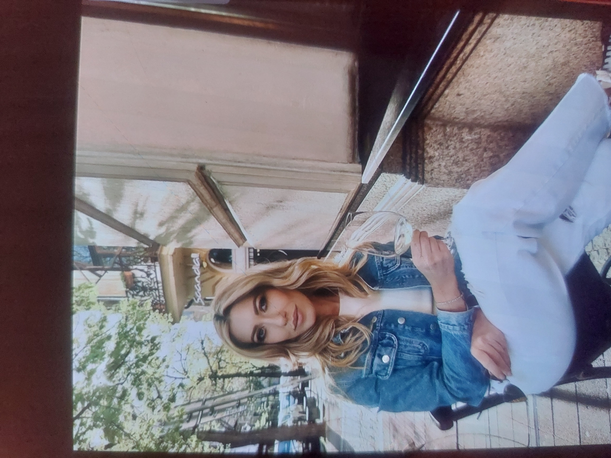 A woman sits at an outdoor cafe in what appears to be Tsarevo, Bulgaria. She wears a denim jacket and white jeans. She holds a wine glass. The cafe appears to be surrounded by trees. The building appears old with stone features.