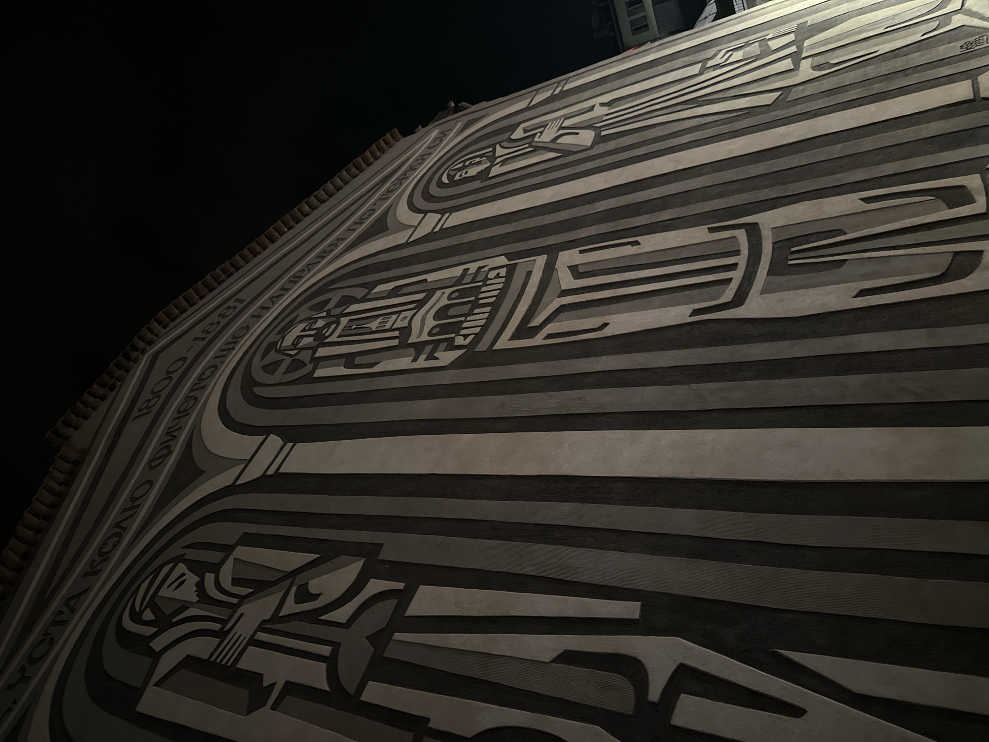 The image shows a large, low-angle shot of a wall mural in Veliko Tarnovo, Bulgaria. The mural depicts stylized figures and machinery in shades of gray and black. The composition is layered with horizontal bands and curvilinear forms. Along the top edge of the mural, fragments of text are visible, including the year "1881." The left side of the image is mostly black, suggesting a nighttime setting. At the upper edge of the image, a few windows are visible. The architecture of the mural shows elements of socialist art and design.