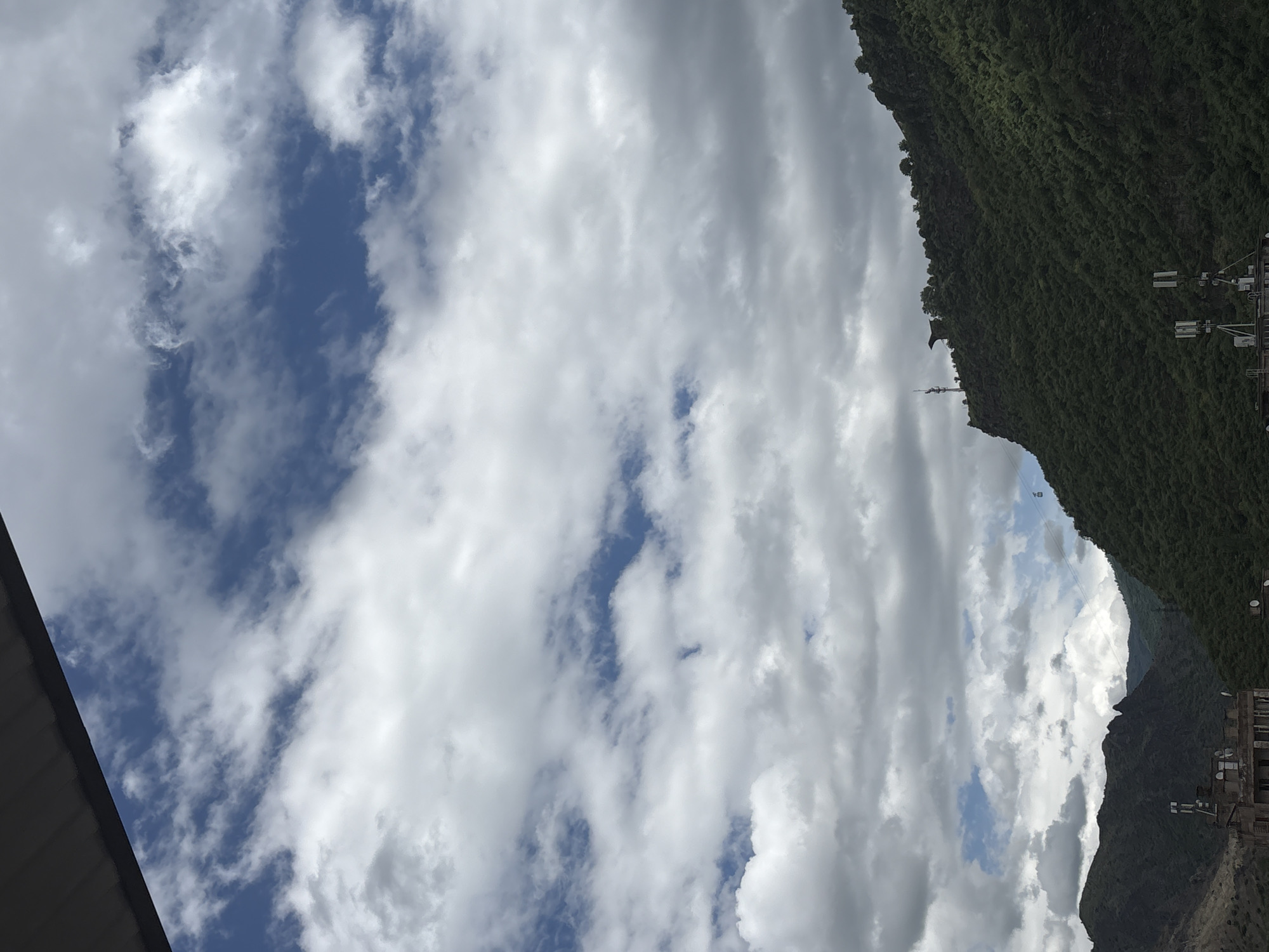 The image presents a partially cloudy sky over a mountainous region. A portion of a building roof is visible in the lower left corner. A densely vegetated mountain slopes up to the right. A communication tower is partially visible on the mountain. The location is Alaverdi, Armenia.