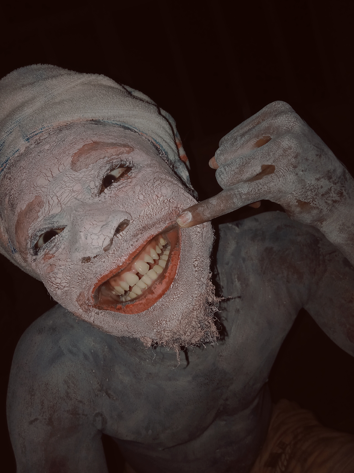 A close-up, dimly lit portrait features a person adorned with extensive body and face paint, set against a dark background. The subject's face is covered in a thick layer of white, powdery paint that has dried and cracked, creating a textured, almost mask-like effect. The lips are painted in a reddish-orange hue and stretched into a wide, open-mouthed smile, prominently displaying a full set of white teeth. One hand, also covered in white paint with visible cracking, points a finger towards the side of the mouth, enhancing the theatrical smile.
The person wears a white, terrycloth-like towel wrapped around their head. Their visible torso and arm are covered in a darker, blue-grey paint, which appears smoother than the facial paint but still shows some texture. The eyes are looking upwards and slightly to the viewer's right, engaging with a lively and expressive gaze. The dark background suggests either a nighttime setting or an indoor space with low lighting. Given the context of Onitsha, Nigeria, this elaborate body art and costume are indicative of cultural performances, masquerades, or traditional ceremonies. The overall impression is one of theatricality and joyful expression. No visible text is present in the image.