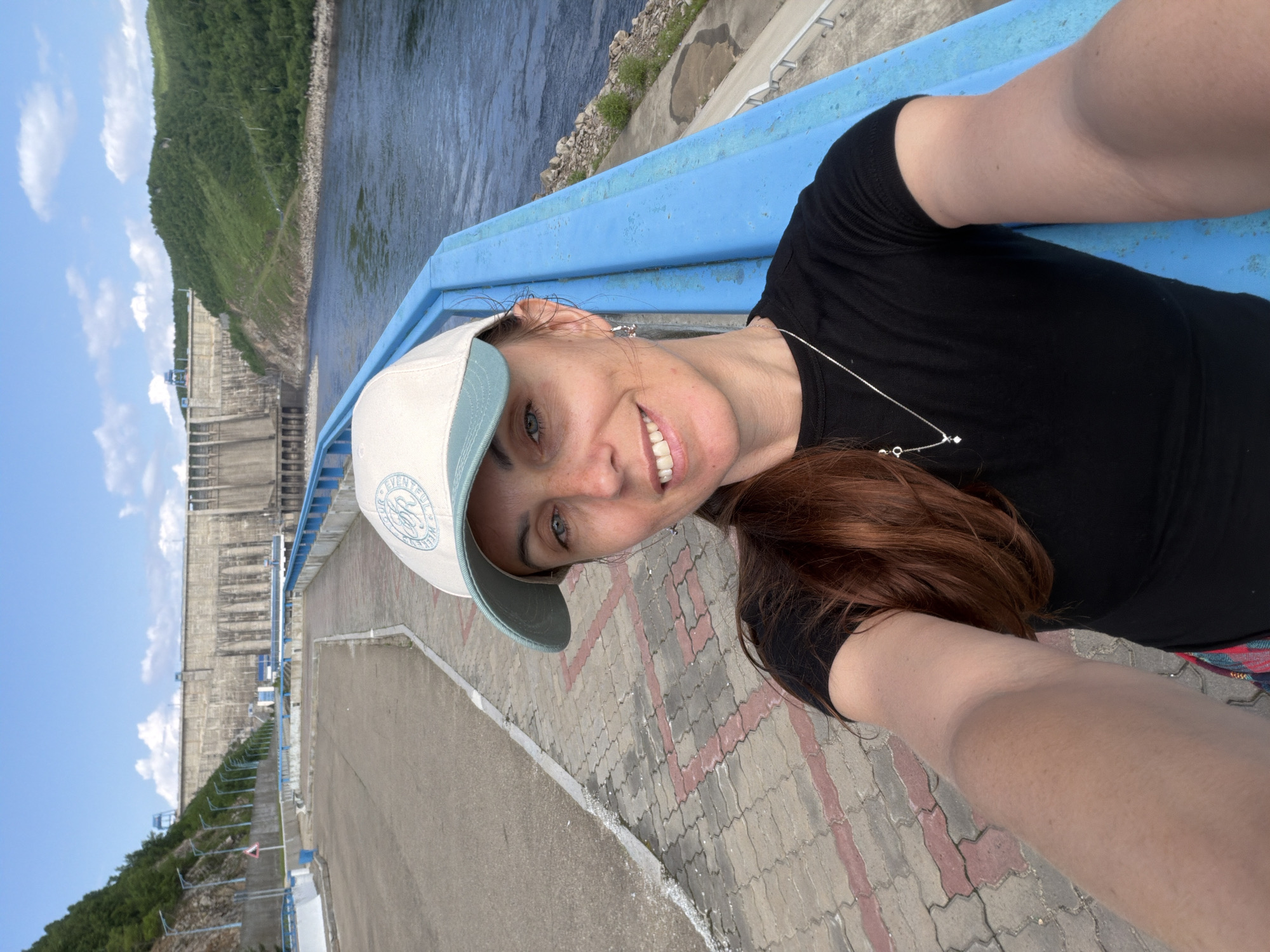 An outdoor, eye-level, medium shot shows a Caucasian woman with medium-length auburn hair, wearing a black t-shirt, a light-colored baseball cap, and a silver necklace. She is smiling and looking at the camera, holding the camera out for a selfie. She is standing on a paved walkway with a blue railing to her right. In the background, a large concrete dam and a wide river are visible under a partly cloudy blue sky. This is the Sayano-Shushenskaya Dam, located in the city of Talakan, Russia.
