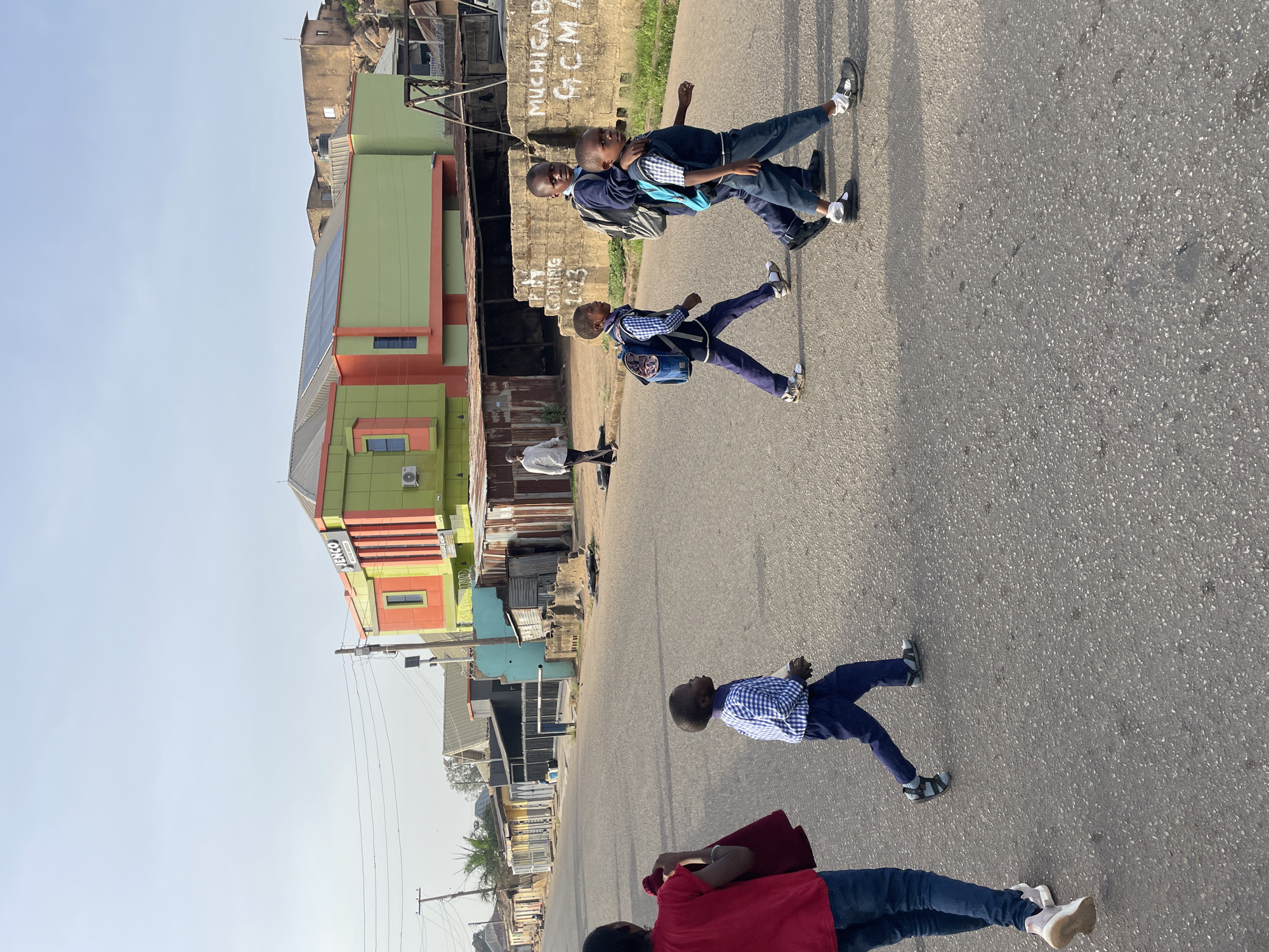 This image captures a street scene in Jos, Nigeria, featuring several children and an adult on a clear, bright day. The perspective is taken from slightly above, looking down a paved asphalt road.
In the mid-ground, three children are walking, two of whom appear to be school-aged and wearing uniforms: dark blue trousers, light blue plaid shirts, and backpacks (one blue, one grey). They are wearing dark shoes and white socks. One child has their arm around another. A third child, slightly ahead of them, wears a blue and white plaid shirt, dark blue trousers, and light sandals.
In the bottom foreground, an individual, possibly an adult or older child, is partially visible from the chest down, wearing a red top, dark blue trousers/jeans, and light-colored sneakers. Another adult figure is visible further down the street on the left, near some buildings.
The street is flanked by various buildings. On the left, a prominent multi-story structure features a vibrant green and orange facade, with solar panels visible on its roof. A sign on this building displays the text "JENCO". Further along the street on both sides, there are other, less well-maintained buildings, some appearing residential, and some constructed with corrugated metal. On the right side of the road, a wall has visible text: "MUCHIGAB GCM A" and "P.H. CONTRAC 2023". Behind these structures, a rocky outcrop with more buildings is visible.
The clear sky and the long shadows cast by the subjects suggest either an early morning or late afternoon setting. Overhead power lines are also visible in the frame. The scene portrays typical daily activity in what appears to be a bustling, semi-urban environment.
