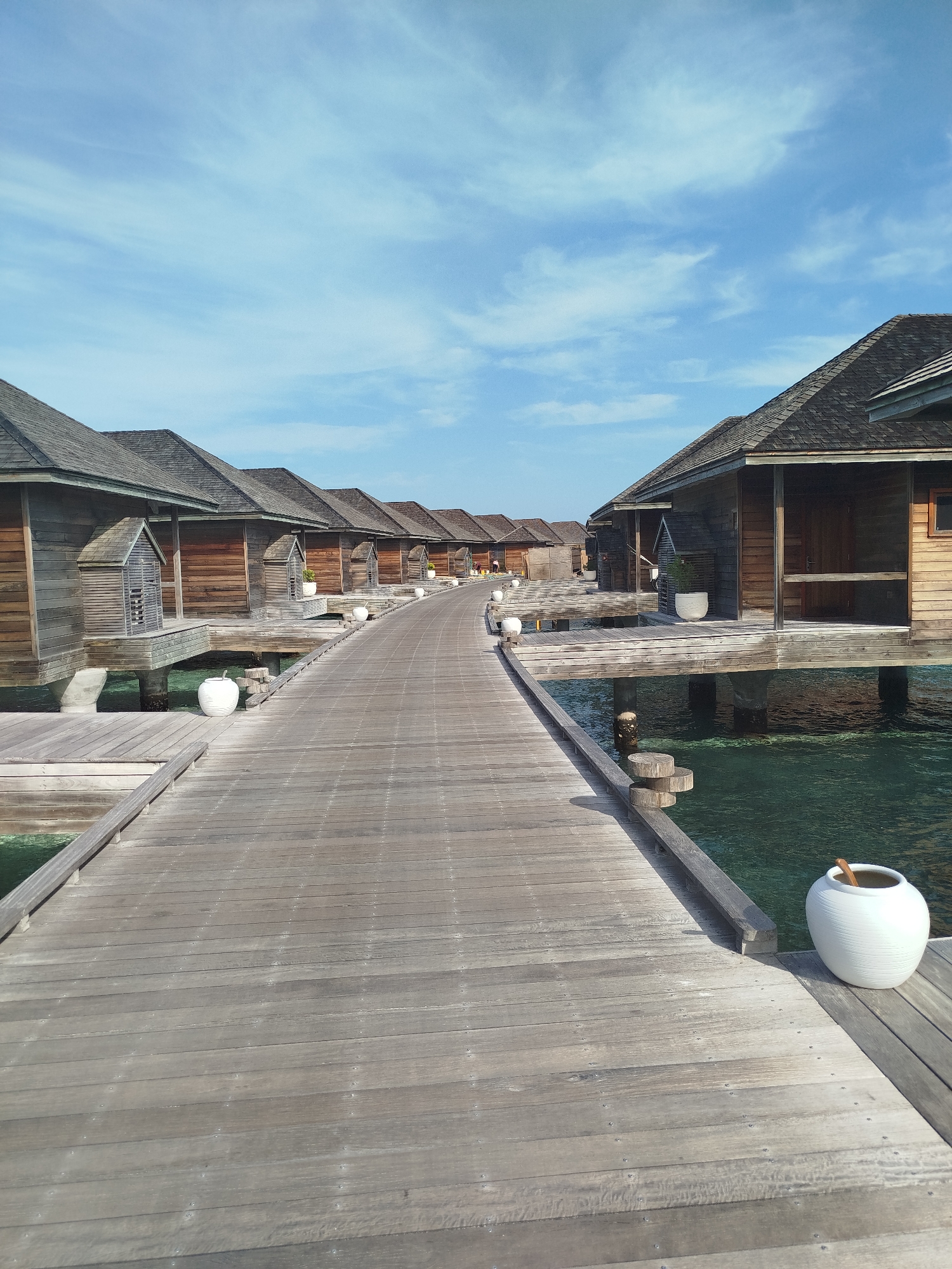 The image depicts a wooden boardwalk leading through a row of overwater bungalows in Hinnavaru, Maldives. The bungalows are constructed of wood with dark-tiled roofs, raised on stilts above the clear turquoise water. The boardwalk is made of weathered wooden planks and runs along the left side of the frame, curving into the distance. White spherical decorative pots are placed periodically along the boardwalk. The sky above is blue with scattered wispy clouds. The scene is serene and captures the typical architecture of a luxury resort in the Maldives. There are no people visible in the image.