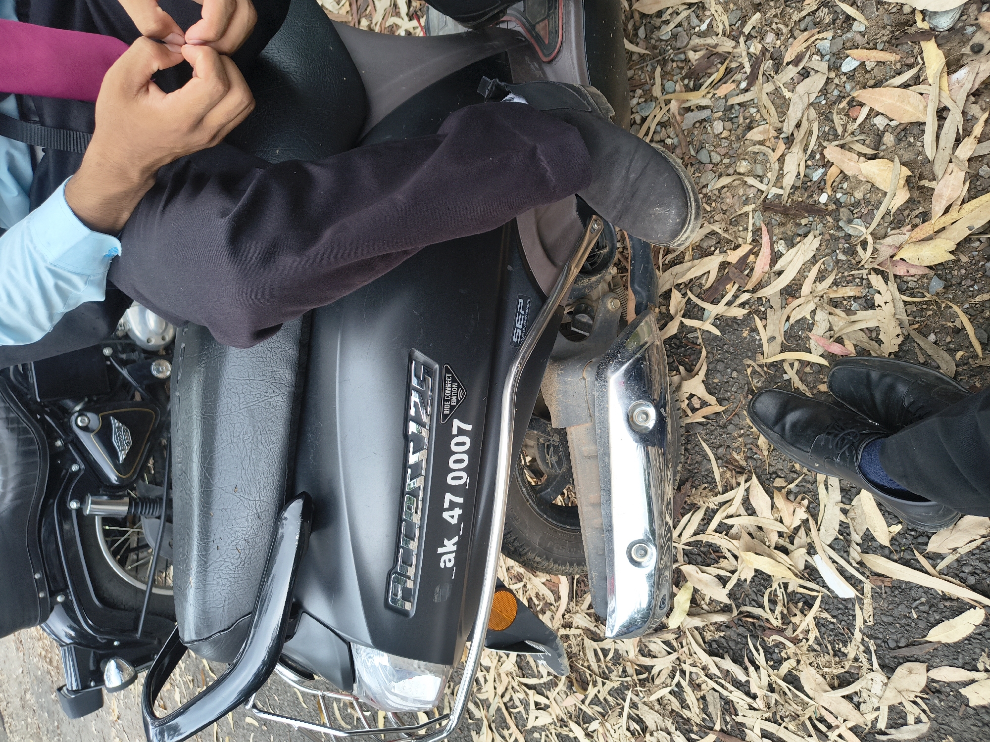 Two individuals are positioned on a dark-colored scooter, with their feet visible in the foreground. The person in the foreground on the left is wearing black formal shoes and dark pants, with blue socks peeking out. The person on the right is wearing black boots and dark pants. A portion of a black motorcycle is visible in the background. The ground is covered with dry leaves and gravel. The scooter has a license plate that reads "ak_47 0007" and a sticker that says "RIDE CONNECT EDITION." The scene appears to be an outdoor urban setting, possibly a street or parking area in Dharamshala, India, given the dry foliage and common vehicle types. The lighting suggests it is daytime.