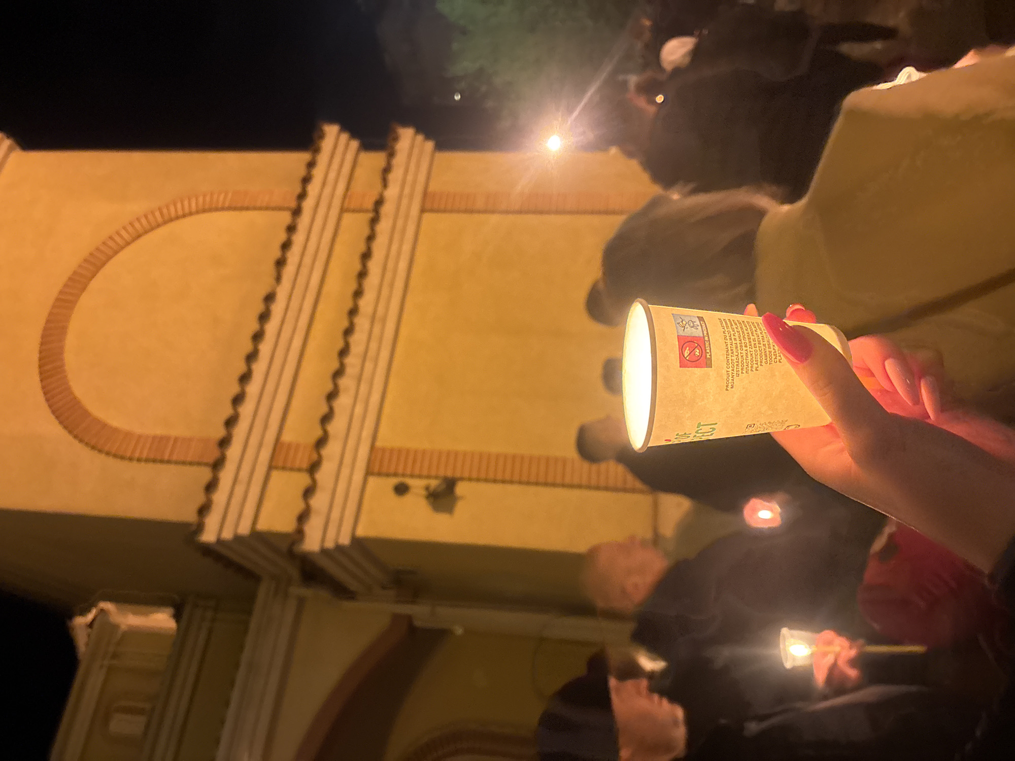 This image shows a candlelit vigil in front of a light-colored building with architectural details, which is likely located in Plovdiv, Bulgaria. A person's hand, with long pink fingernails, holds a candle in a white paper cup, prominently in the foreground. The cup has text printed on it, seemingly related to plastic content in the product. In the background, a crowd of people holds similar candles, their faces illuminated by the soft glow. There is a dark sky overhead, with a bright light source, possibly a streetlight. The architecture appears to have a brick-detailed arch and pilasters. The building's light color and design suggest an older structure, possibly a monument or a historical building. The setting implies a solemn or commemorative event.