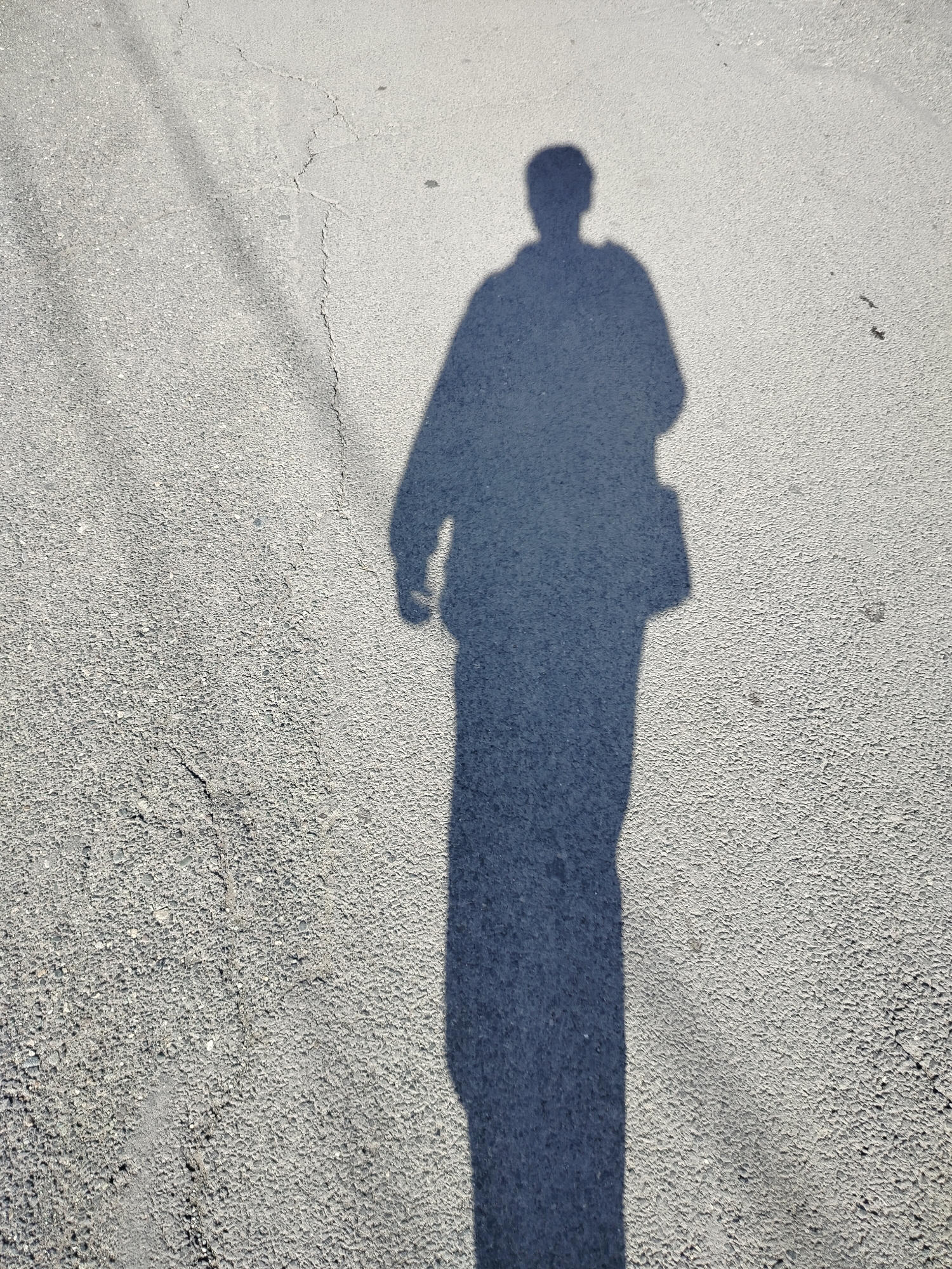 The image shows the shadow of a person on a gray asphalt surface, likely a street or sidewalk. The person appears to be wearing a jacket and carrying a bag on their right side. The shadow indicates that the sun is positioned to the side and slightly behind the person. The surface has some cracks and variations in texture. There is no other information in the image that would indicate the location. Therefore, it's not possible to pinpoint whether this image was taken in Yerevan, Armenia, or any other specific city.