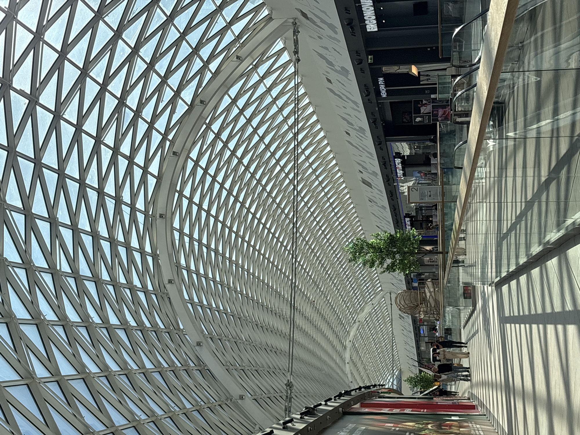 The image shows the interior of the Galleria shopping mall in Perm, Russia. The mall features a large, arched glass roof with a geometric pattern. Below the roof, there are several levels of retail stores, visible as dark shapes with some signage. A walkway runs along the right side of the image, with glass railings casting shadows on the floor. Groups of people are walking along the walkway. There are also decorative trees and sculptural elements placed within the mall.