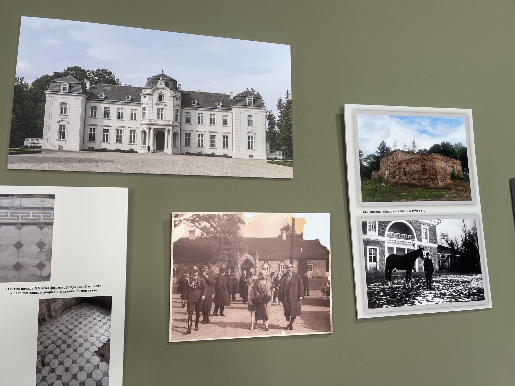 The image displays an exhibition of photographs mounted on a green wall. The photographs feature architectural elements and historical scenes.
The largest photograph at the top left shows a grand white manor house with a distinctive mansard roof and ornate detailing. It is set against a backdrop of trees and a cloudy sky. A person is visible near the entrance of the manor.
To the right of the manor house photograph is a smaller framed photograph of a dilapidated stone building with a partially collapsed roof, surrounded by overgrown vegetation. This photograph is captioned with "Канюшенная официна сейчас и в 1930-х гг." which translates to "Stable office now and in the 1930s."
Below the manor house photograph, on the left, is a vertical photograph featuring two images. The top image shows a tiled floor with a geometric pattern. The bottom image also depicts a tiled floor, but this one appears to be in a state of disrepair, with exposed earth and broken tiles. This section is accompanied by Russian text that reads "Плитка начала XX века фирмы Дзевульский и Ланге в главном здании дворца и в «замке Тизенгауза»" which translates to "Tile from the early 20th century from the Dziewulski and Lange firm in the main palace building and in the 'Tyszkiewicz Castle'."
The largest photograph in the bottom half of the image is a sepia-toned historical photo showing a group of formally dressed people walking outdoors in front of a building. One man in the foreground is wearing a military-style uniform.
Below the historical group photo, on the right side, is another framed photograph. This black and white image shows a horse standing next to a person in uniform, in what appears to be a courtyard in front of a building with arched features.
The overall impression is of an exhibition dedicated to the history and architecture of a specific estate, likely in the Жалудоцкі сельскі Савет region of Belarus, showcasing both its past grandeur and its present state.