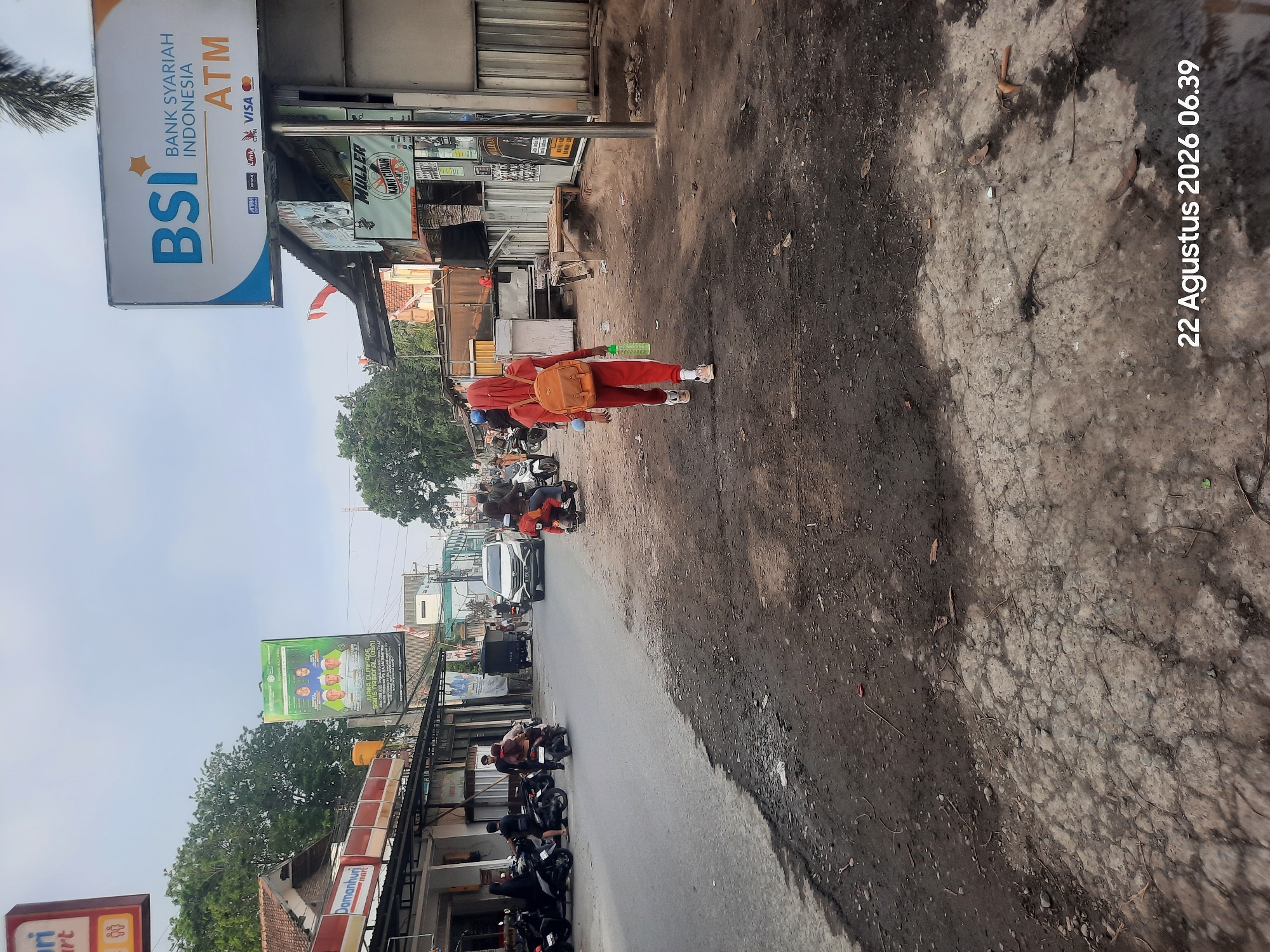 The image shows a street scene in Pajarakan Kulon, Indonesia, on August 22, 2026, at 06:39. A young woman wearing a red hijab and a matching red tracksuit, carrying an orange backpack and a green water bottle, walks away from the camera down a dusty street. To her left, a white car is parked on the side of the road next to several motorcycles. In the background, various buildings line the street, including a storefront with a sign reading "Damanhuri mart" and a large billboard advertising "BSI BANK SYARIAH INDONESIA ATM". A traffic light can be seen hanging over the street. The overall environment appears to be a modest urban or suburban area with a mix of commercial and residential structures.