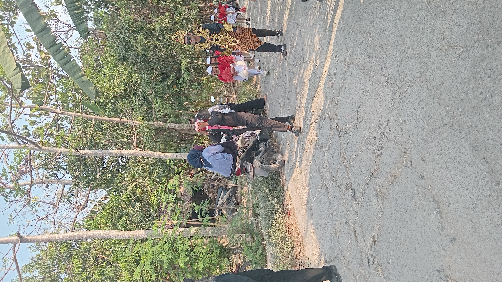 The image captures a procession on a paved road in Kedungwaru, Indonesia. A group of children in red and white attire, some holding what appear to be fans, walk in the distance. A young man in elaborate gold and brown traditional clothing, with sunglasses and a headdress, walks prominently in the foreground. To his left, a person in a black outfit with a red sash holds a decorative headdress. A woman wearing a blue headscarf and shirt is seated on a motorcycle to the left of the road. The background consists of lush green trees and vegetation, with a clear sky overhead. The road surface is cracked and uneven.