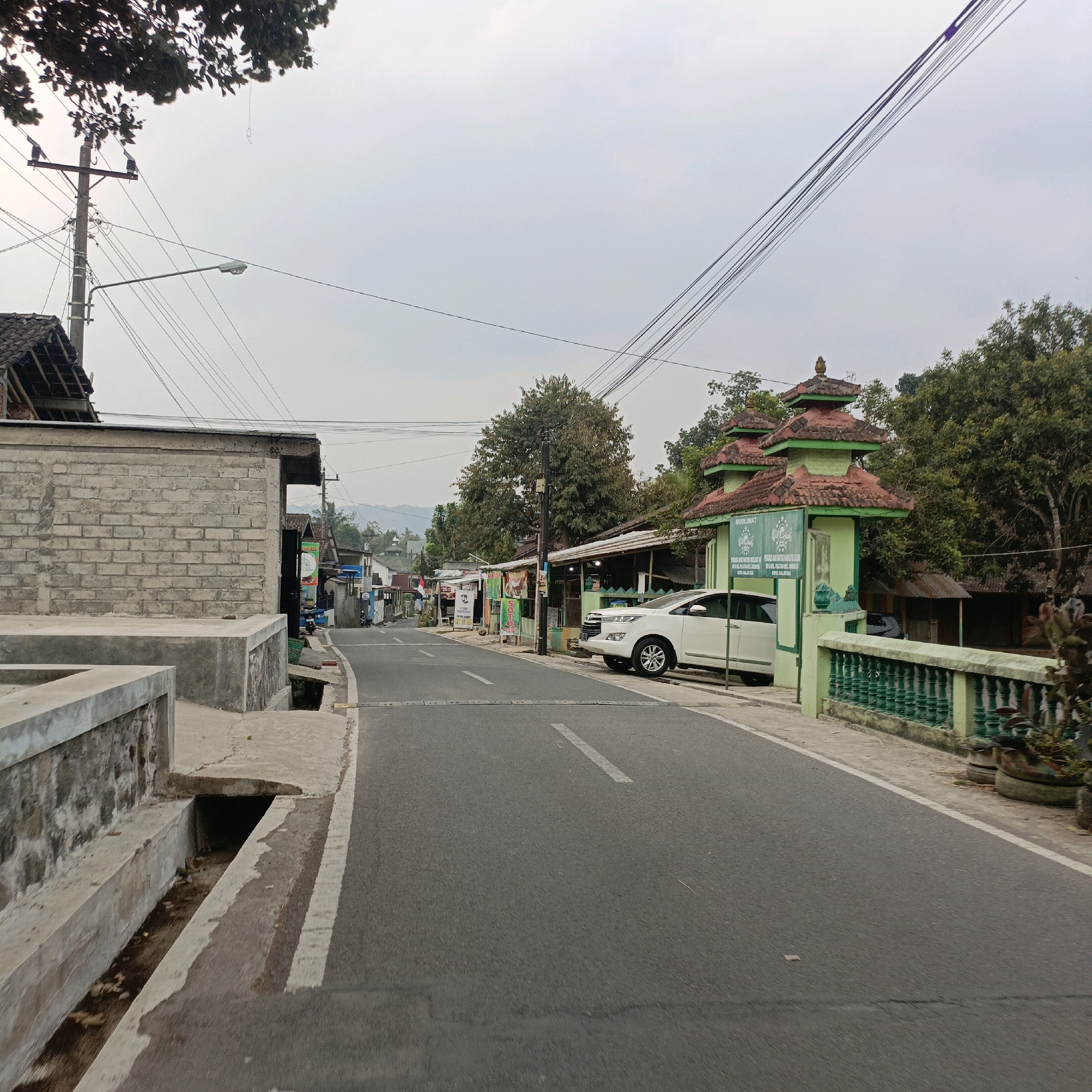 The image depicts a street scene in Salatiga, Indonesia, characterized by a gray, overcast sky. The road, paved with asphalt, features white dashed lines and a speed bump. On the right side of the road is a light green building with a traditional Indonesian tiered roof, marked with the word "MUSLIMAT" in green lettering and an emblem. A white SUV is parked in front of this building. Adjacent to the building is a decorative green fence. On the left side of the road, a partially constructed gray brick building with a concrete foundation and drainage ditch is visible. Further down the road, a row of small shops and stalls line the street, with trees and other vegetation scattered throughout the scene. Utility poles with numerous electrical wires are prominent in the background. The overall atmosphere suggests a quiet, somewhat rural or suburban environment.