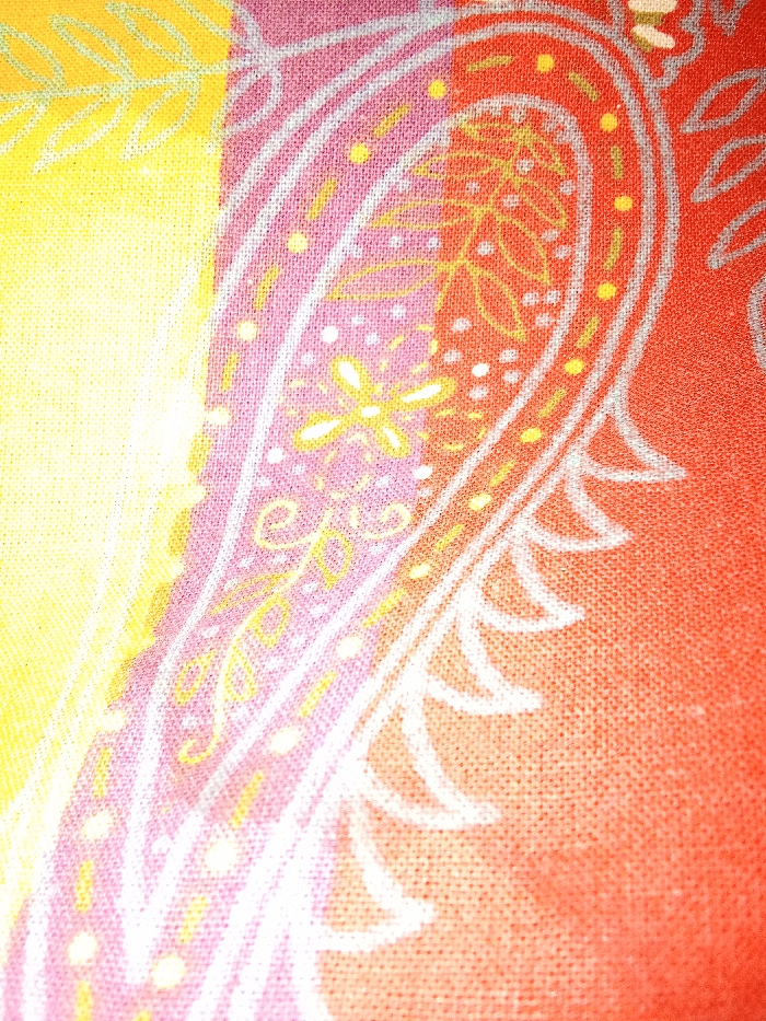 This image displays an extreme close-up of a vibrantly patterned textile fabric, located in the city of Yarun, Ukraine. The fabric features a prominent paisley design outlined in white. The background transitions across vertical color blocks: a bright yellow on the left, merging into a pinkish-lavender hue in the central area encompassing a portion of the paisley, and then shifting to a reddish-orange on the right. Within the white paisley contour, intricate details are present, including small yellow floral motifs with white centers, scattered yellow and orange dots, and delicate yellow curvilinear and foliate embellishments. The woven texture of the fabric is clearly discernible across the entire surface.