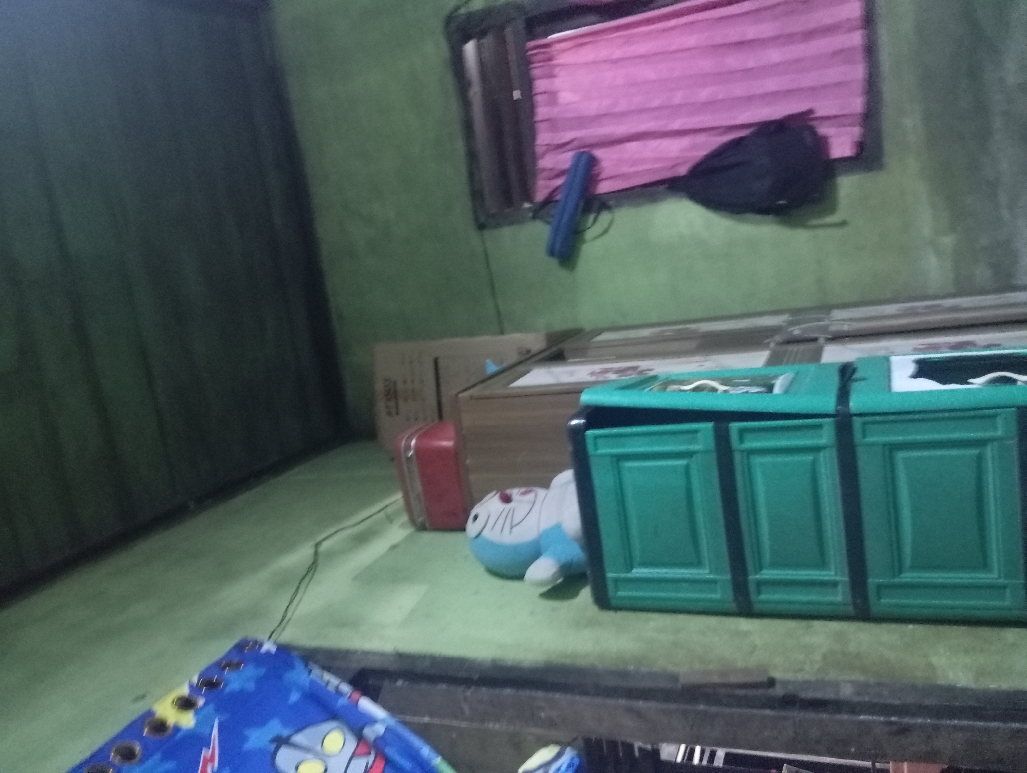 The image depicts an indoor scene, likely a room in Rejosari, Indonesia, characterized by its simple, functional furnishings. The walls are painted a muted green, and the ceiling appears to be made of dark, horizontal wooden planks.
Dominating the left side of the frame is a tall, green plastic cabinet with three visible drawers, each featuring a recessed rectangular panel design. Perched on top of this cabinet is a light blue plush toy resembling the character Doraemon. Behind the Doraemon toy, on a higher shelf, is a red rectangular container. Further back, a large cardboard box with some text is visible.
To the right of the cabinet, a window is covered by a bright pink curtain, drawn to one side. A black backpack is hanging on the wall to the left of the window. Above the window, a blue cylindrical object is mounted horizontally on the wall.
In the upper left corner of the image, a portion of a blue curtain or tapestry with a cartoon character (possibly Ultraman) is visible, hung with grommets.
The overall impression is one of a lived-in space, sparsely decorated and utilitarian. The lighting is dim, contributing to the slightly shadowy atmosphere of the room. The photograph is taken from a slightly low angle, looking upwards towards the ceiling and window.
