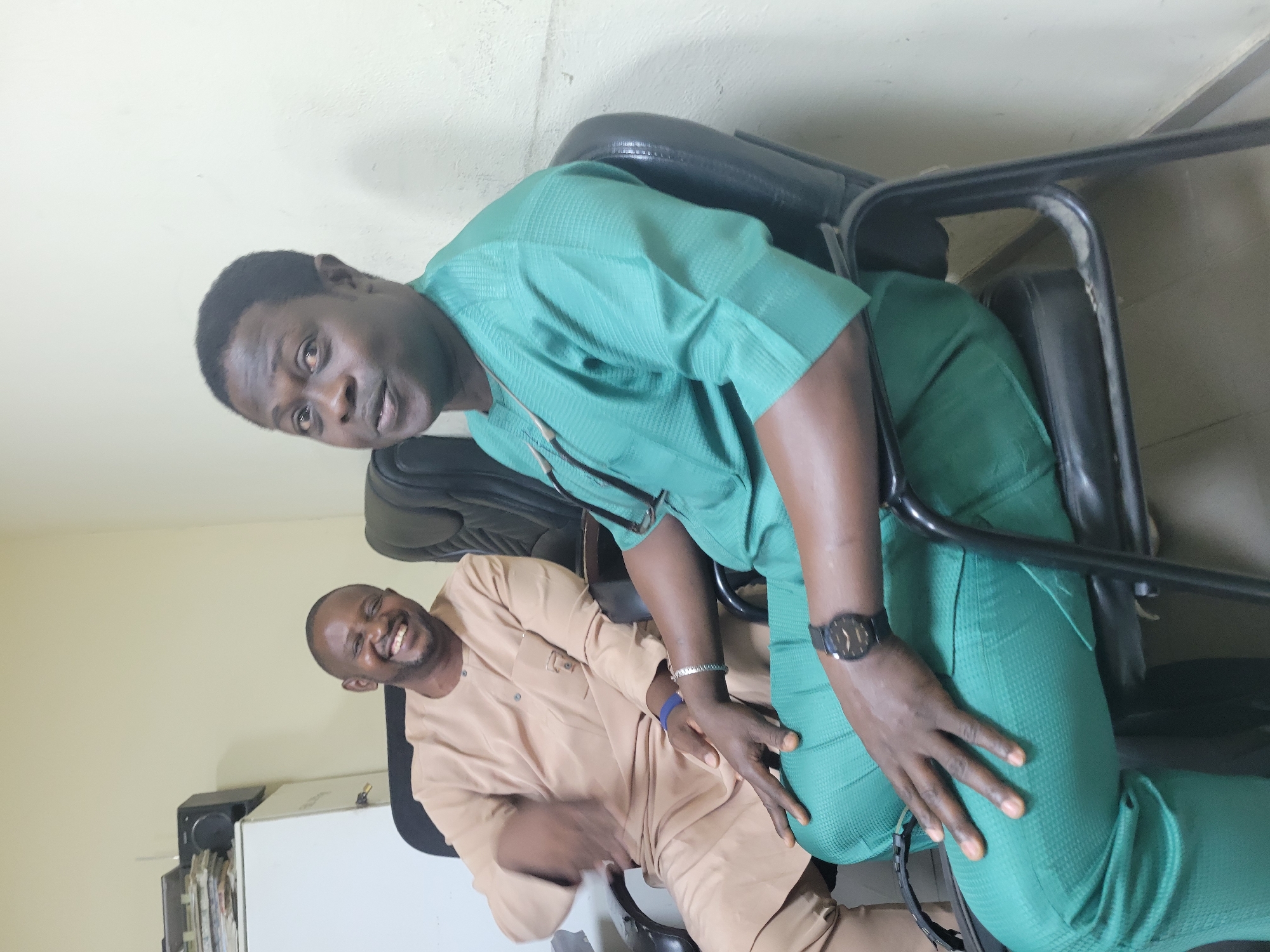Two men are seated in office chairs in what appears to be an office environment. The man on the left is wearing a light brown traditional outfit and is laughing, with his right arm extended. The man on the right is wearing a green traditional outfit and is looking towards the camera. He has a watch on his left wrist. The room has pale walls and a filing cabinet is visible in the background. The image location is not specified beyond being in Abuja, Nigeria.