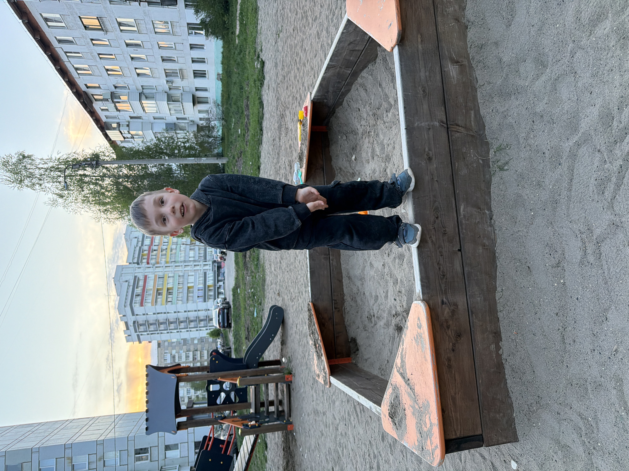 A child stands on the edge of a wooden sandbox filled with sand at a playground in Chaikovsky, Russia. The child wears a dark jacket and pants with blue shoes. In the background are several multi-story apartment buildings and playground equipment, including a slide. The image is shot outdoors in what appears to be late afternoon or early evening, judging by the light.