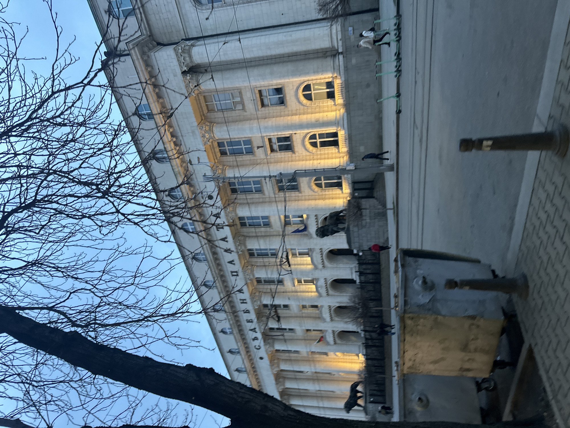 The image shows the Largo in Sofia, Bulgaria, specifically the National Assembly building. The building is a large, white structure with multiple floors and arched windows. Cyrillic letters spell out "СЪБРАНИЕ НА ПАЛАДИ" across the front of the building. A row of bare trees is visible on the left side of the image. People are walking in front of the building. Modern scooters are parked along the edge of the street. A metallic, possibly temporary structure is positioned on the right side of the sidewalk. The sky is overcast.