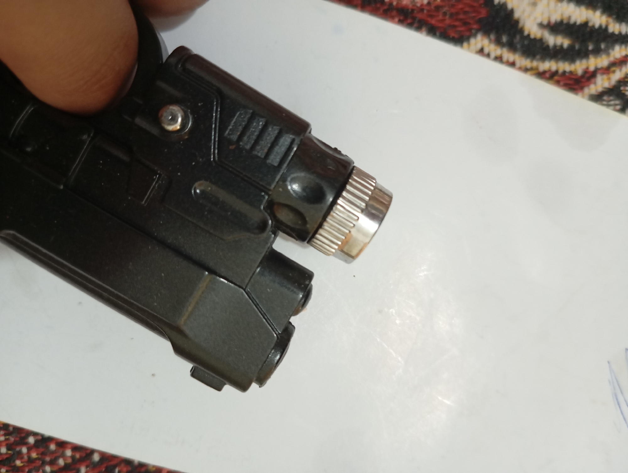 A close-up, overhead shot shows a black, gun-shaped lighter being held by a person's thumb on the left side of the frame. The lighter is positioned diagonally, with its nozzle pointing towards the right and slightly upwards. The nozzle is metallic and ridged. The surface beneath the lighter is white and appears to be paper, with some faint markings or smudges. In the background, a patterned fabric with red, black, and yellow colors is visible. The lighting is bright, creating some reflections on the lighter and the white surface.