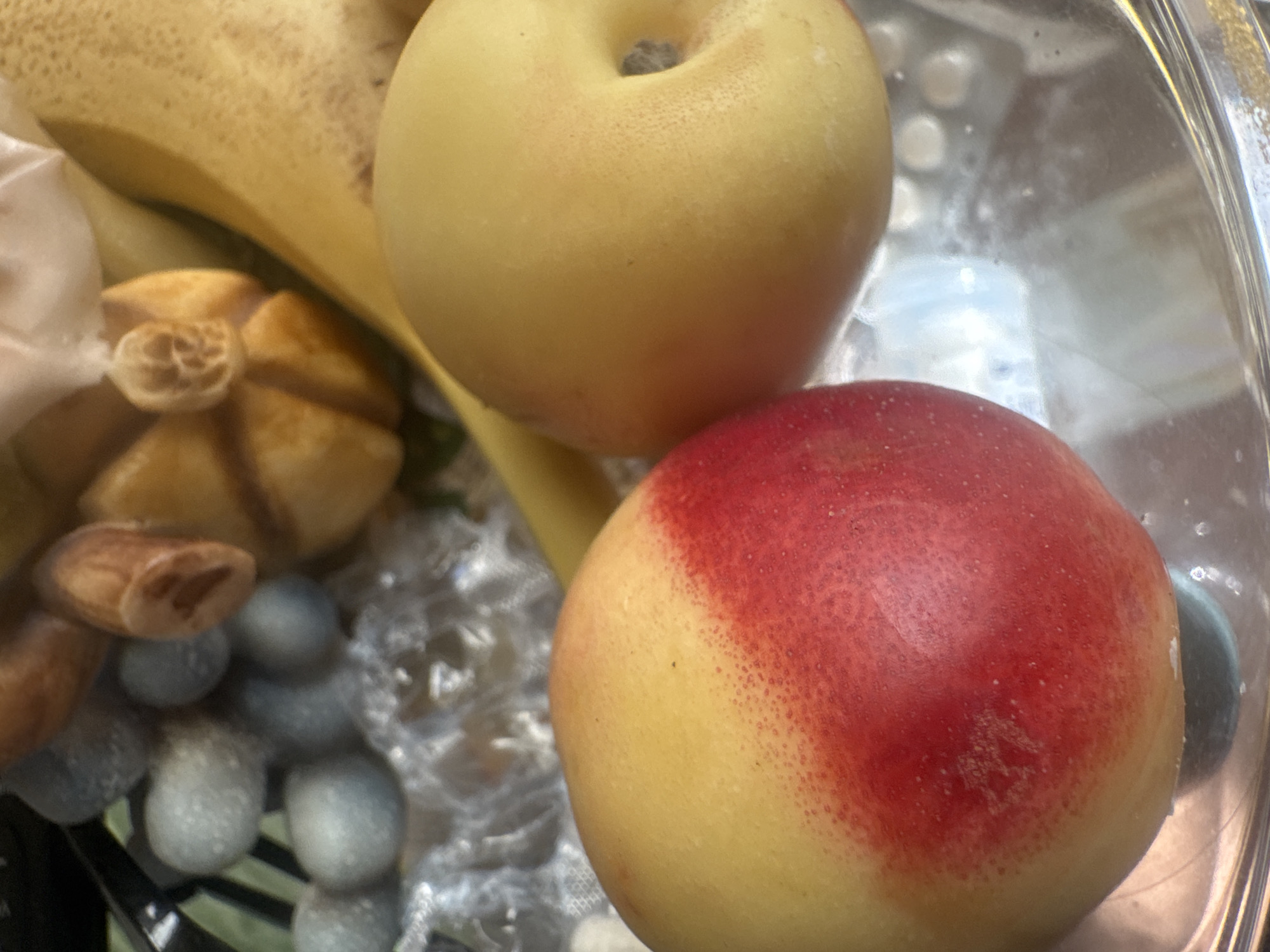 This is a close-up, high-angle shot of fruit in what appears to be a transparent bowl. In the foreground, a red and yellow nectarine or peach is visible on the left, with its skin showing a transition from yellow to a vibrant red. To its right is a yellow apple with a dimple. In the background, out of focus, are what seem to be light blue grapes and a portion of a banana. Some textured plastic wrap is also visible between the fruits. The scene is brightly lit, suggesting it is indoors or under artificial light. There is no indication of the city of Sochi, Russia, or any other specific location in the image.