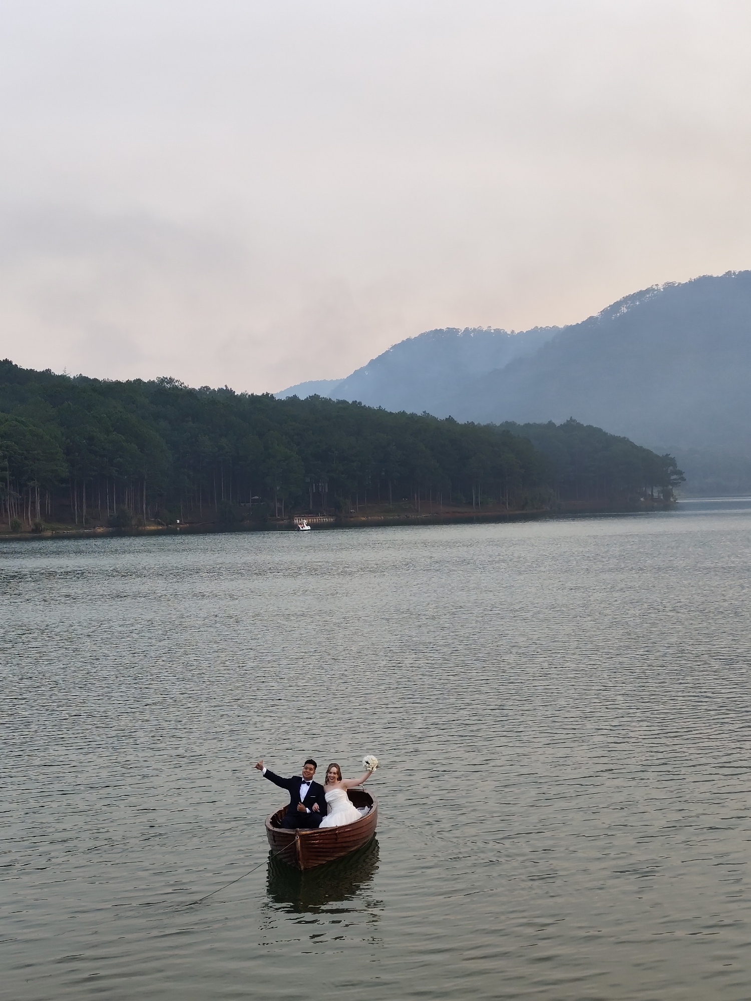 A long outdoor shot of a wedding couple in a small wooden boat on a lake, presumably Xuan Huong Lake in Da Lat, Vietnam, given the surrounding pine forest and distant mountains under a muted sky. The bride, on the right, is dressed in a white wedding gown and holds a bouquet. The groom, on the left, wears a tuxedo and raises his left arm in a celebratory gesture. They are the focal point of the image, positioned in the boat in the lower center of the frame. The lake surface is calm, with subtle ripples visible. The forested shoreline is visible in the background, transitioning into hazy mountains. The sky is overcast, contributing to the overall soft lighting and muted colors of the image.
