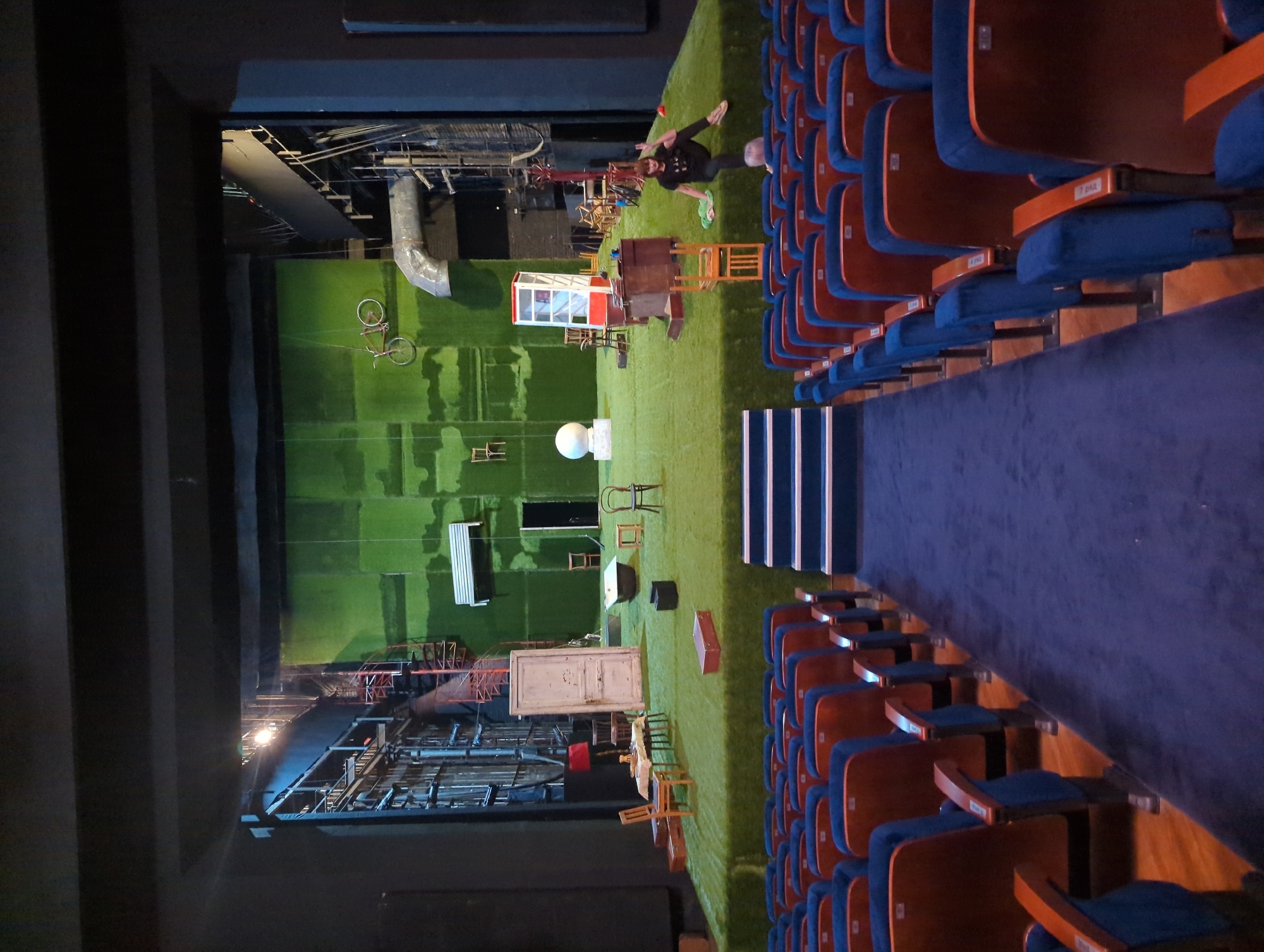 The image depicts a view of a theatre's stage from the audience's perspective. The stage is covered in green material resembling grass, and various props are scattered around, including chairs, tables, a bicycle hanging from above, a phone booth, and other miscellaneous objects. A person is lying on the stage near the edge. The theatre seats are visible on the right side of the image, upholstered in blue and arranged in rows. The lighting is a combination of stage lights and ambient light from the theatre. The photograph's location cannot be determined from the image alone.
