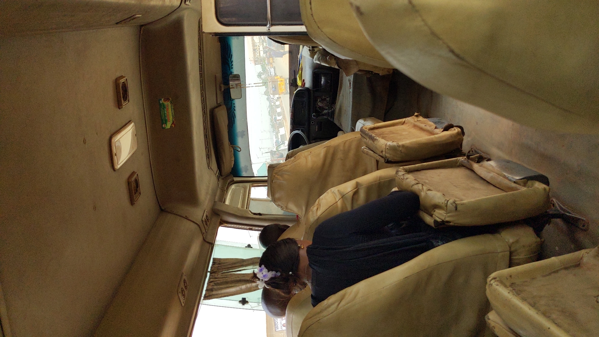 This media file captures the interior of what appears to be a passenger van or small bus, looking forward towards the driver's area and front windshield. The image is rotated, showing the roof on the left and the floor on the right.
**Subjects:**
In the foreground, a person is seated with their back to the camera, wearing a dark top. Their hair is tied back, adorned with a light-colored, possibly purple or white, floral accessory. Another person's head is partially visible beside them. The vehicle is equipped with multiple rows of passenger seats, upholstered in a light beige or tan material. These seats appear well-used, with visible wear, tears, and exposed structures on some cushions and seatbacks. The ceiling and interior side panels are a similar light color. The driver's dashboard, steering wheel, and rearview mirror are visible through the front windshield.
**Activity:**
The vehicle seems to be stationary, with passengers inside. The relaxed posture of the visible passenger suggests they are either waiting to depart, having just arrived, or paused during a journey.
**Notable Details:**
The lighting indicates it is daytime, with bright light coming through the windows and windshield. The exterior view through the front window reveals a dusty environment with other vehicles parked, suggesting a roadside stop, a motor park, or a busy street. The front windshield has a blue decorative tint at the top. Curtains are pulled back on the side windows. The interior condition, with worn seating, suggests a vehicle used for regular public transport. The context of Ado, Nigeria, aligns with the appearance of a typical regional transport vehicle.
**Visible Text:**
On the overhead panel, a small green and red sticker or air freshener is visible, partially reading "Sunshine" or a similar brand name.