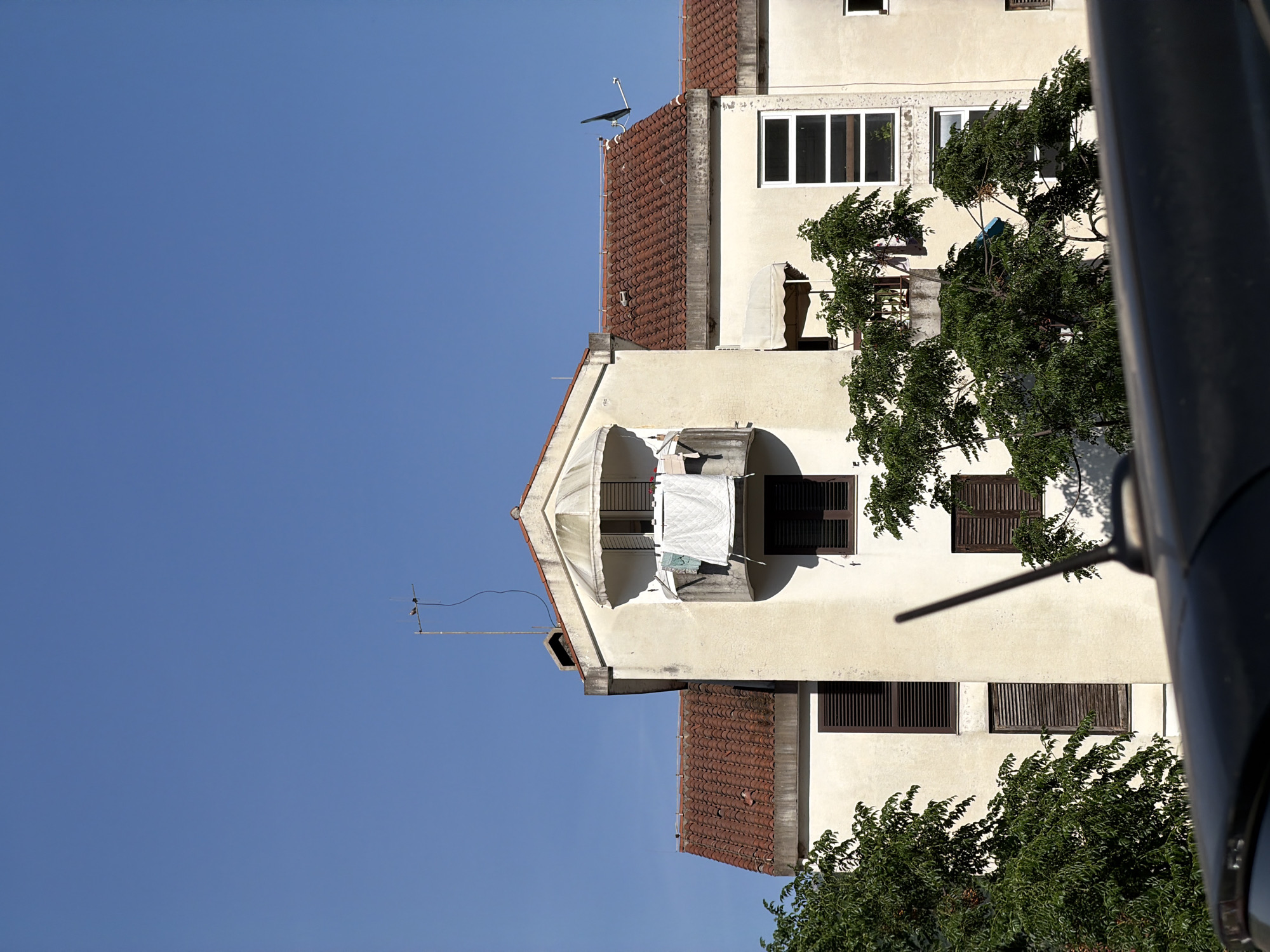 The image shows a multi-story building in Budva, Montenegro. The building has a cream-colored facade with dark brown shutters. Some laundry hangs from a balcony. Trees obscure portions of the building. The sky is clear and blue. An antenna is visible on the roof. A portion of a dark, cylindrical object is visible in the foreground.