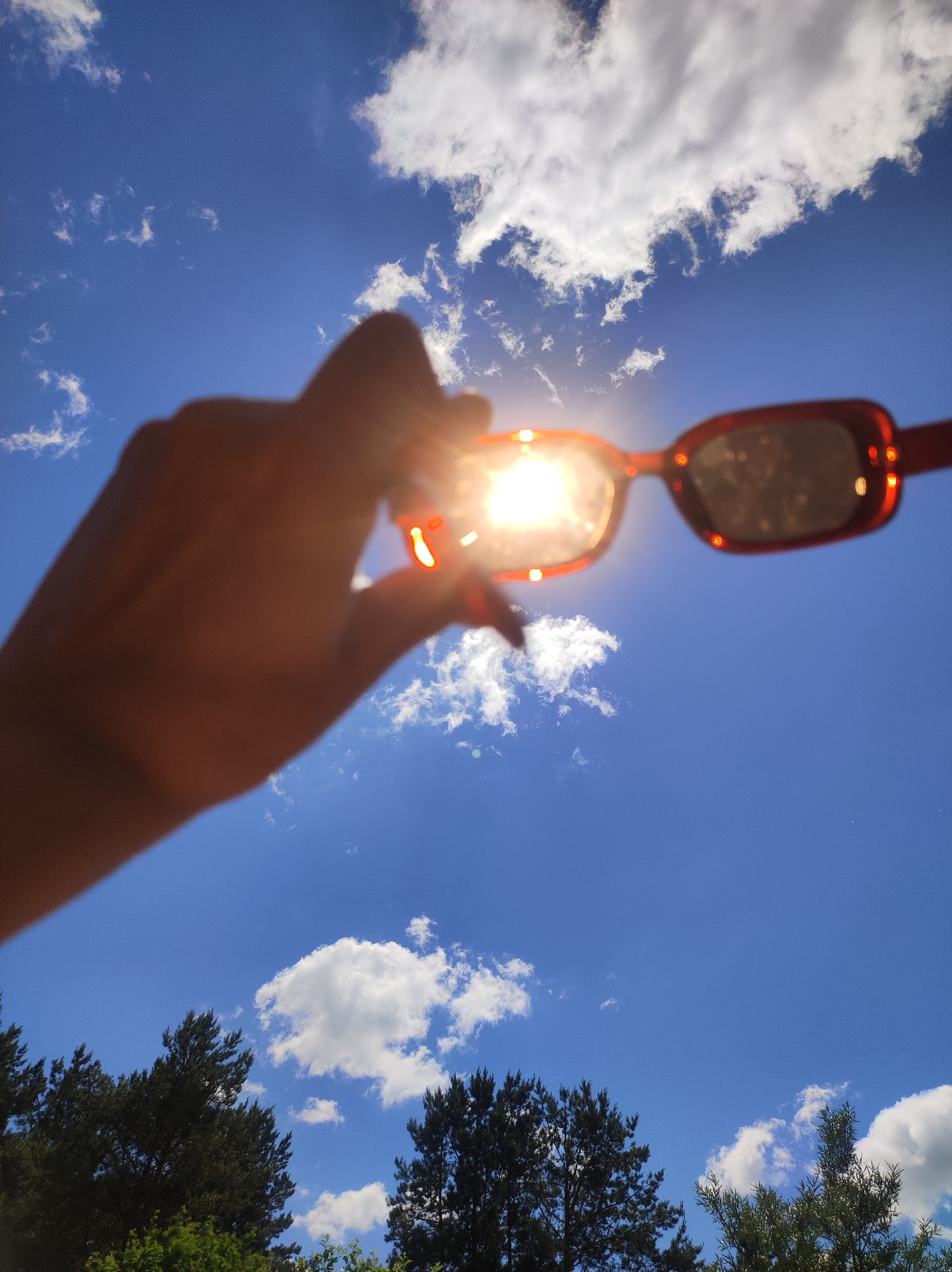 A person's hand, partially visible and out of focus, holds a pair of orange-rimmed sunglasses up to the bright sun, creating a lens flare. The sunglasses are positioned in the foreground, obscuring some of the sun's glare. The background is a bright blue sky with scattered white clouds. Below the sky, the tops of dark green pine trees are visible, suggesting a natural or park-like setting. The overall image captures a sunny day, likely outdoors. No specific location in the city of Фоки, Russia, can be determined from the image alone.